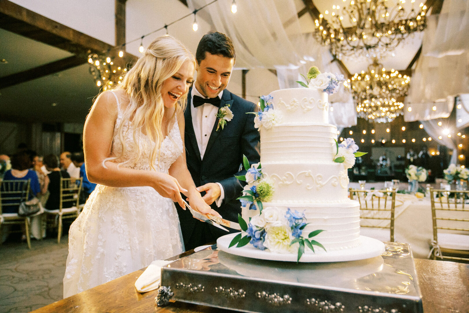 At a Normandy Farm wedding, the bride and groom are smiling as they cut a large, white wedding cake adorned with blue and green flowers, surrounded by the elegance of chandeliers in the reception hall.