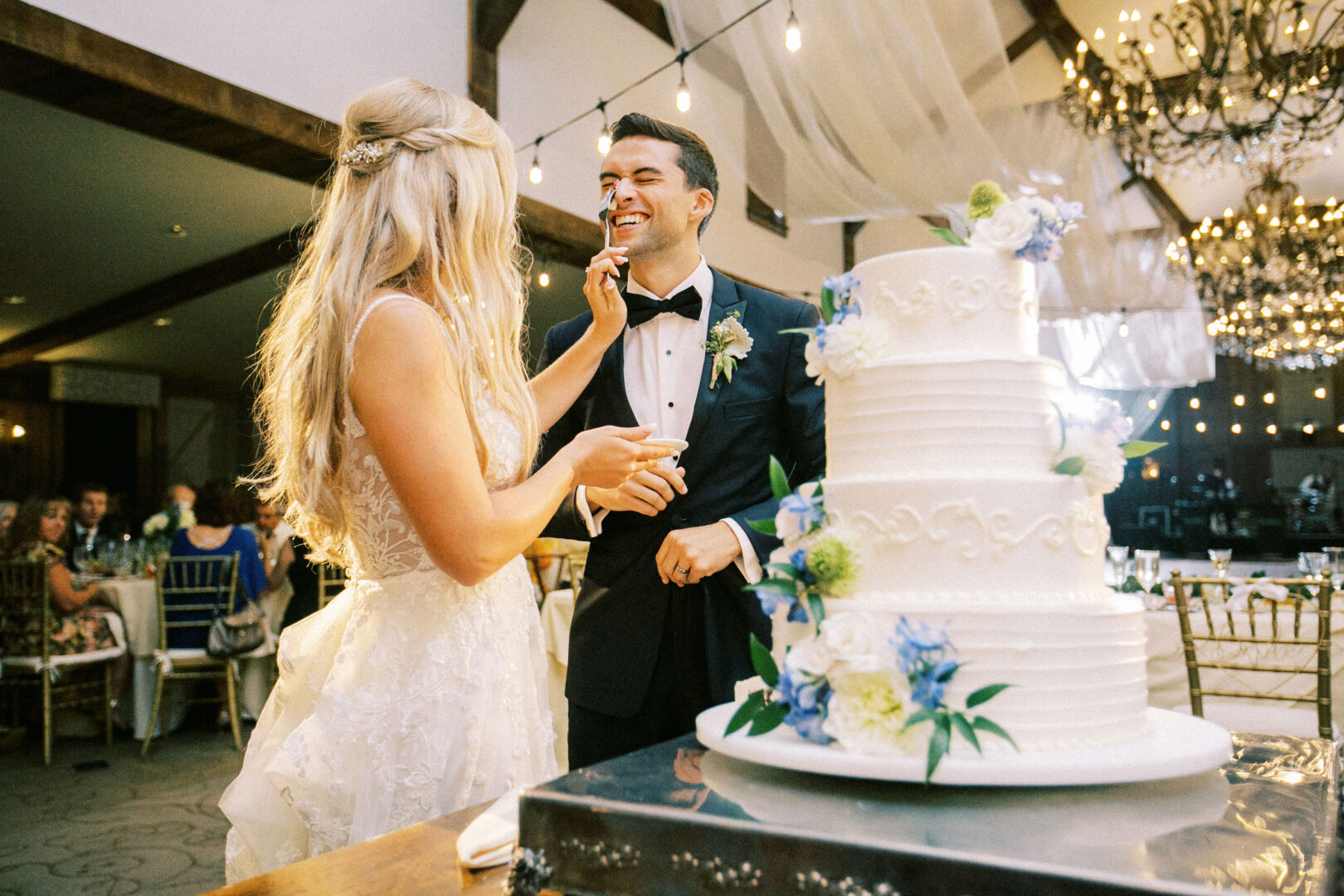 At a Normandy Farm wedding, the bride and groom in formal attire share a tender moment as he gently touches her face with frosting from the wedding cake in an elegantly decorated indoor venue.