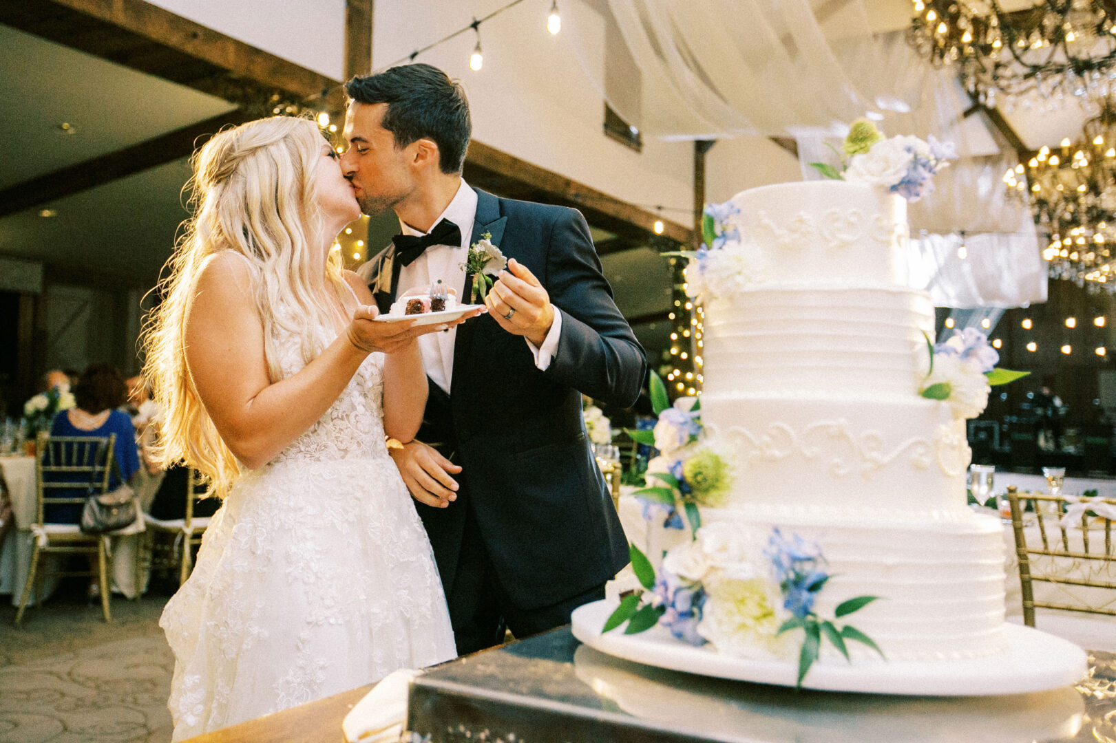 At their Normandy Farm Wedding, the bride and groom share a sweet kiss while holding a plate of cake slices, with the beautifully decorated tiered cake standing proudly in the foreground.