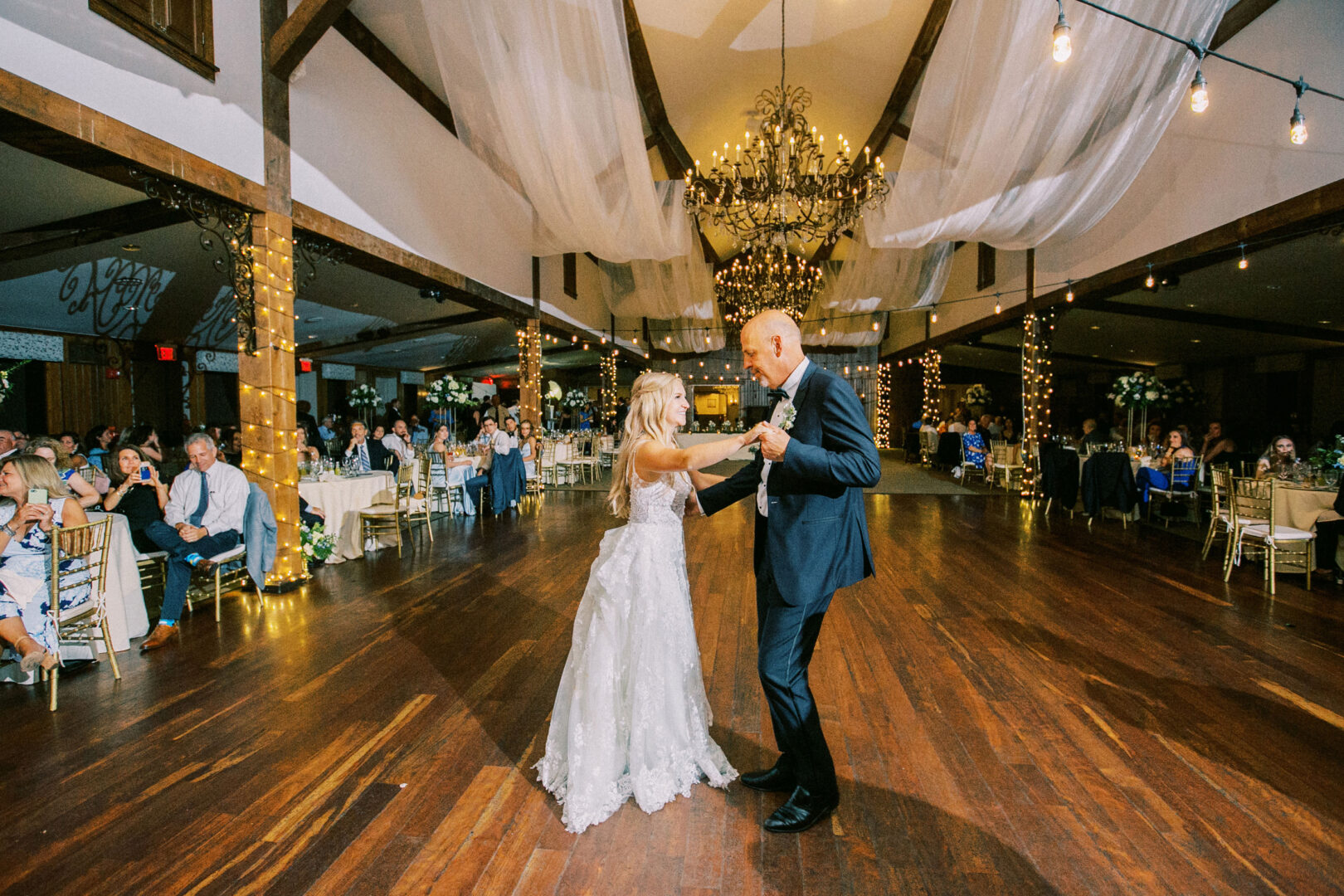 In the beautifully adorned reception hall of Normandy Farm, a bride and groom elegantly dance as guests watch from tables in the background.