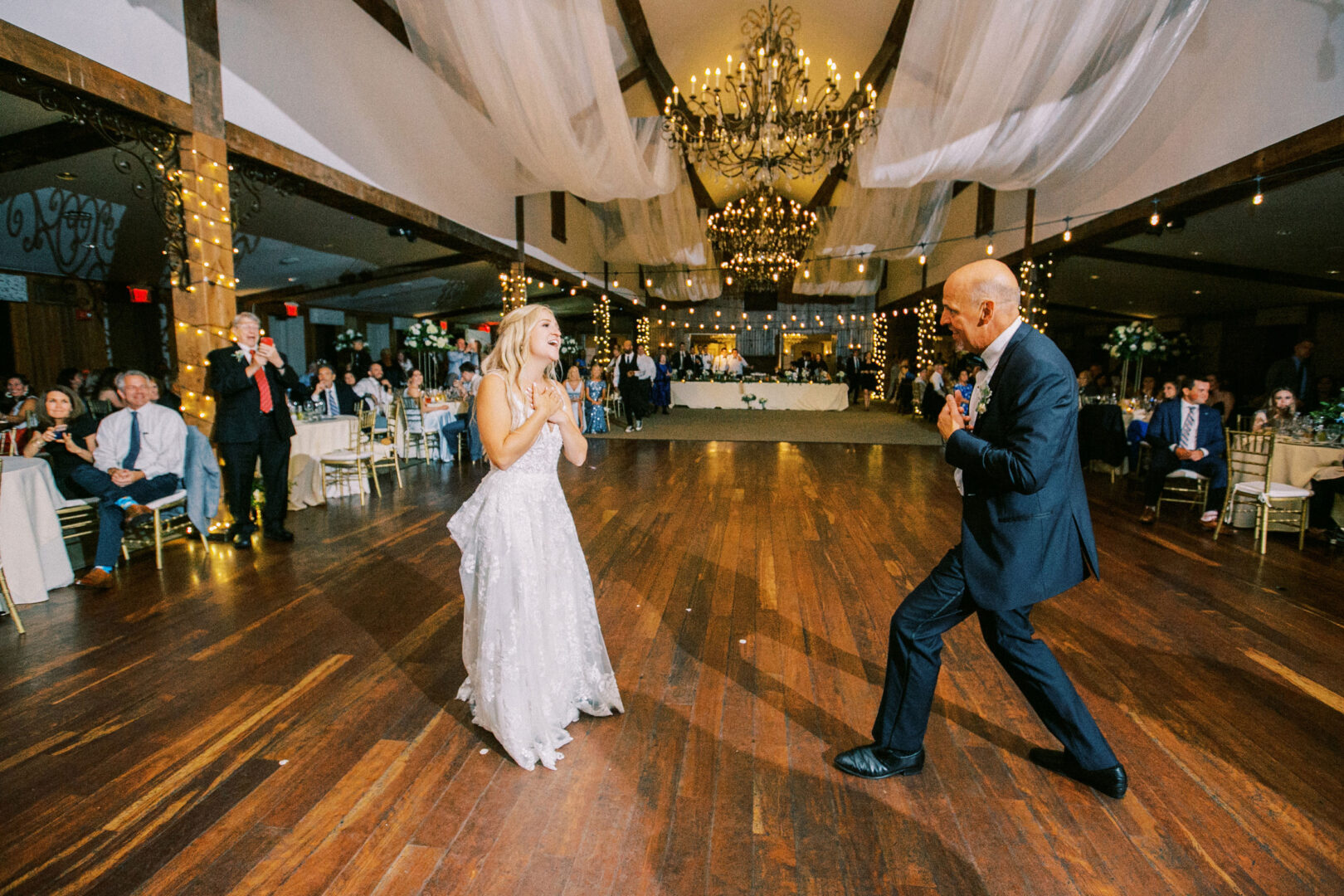 At the Normandy Farm Wedding, a bride and an older man dance joyfully on a wooden floor. Guests seated around them smile as chandeliers illuminate the celebration.