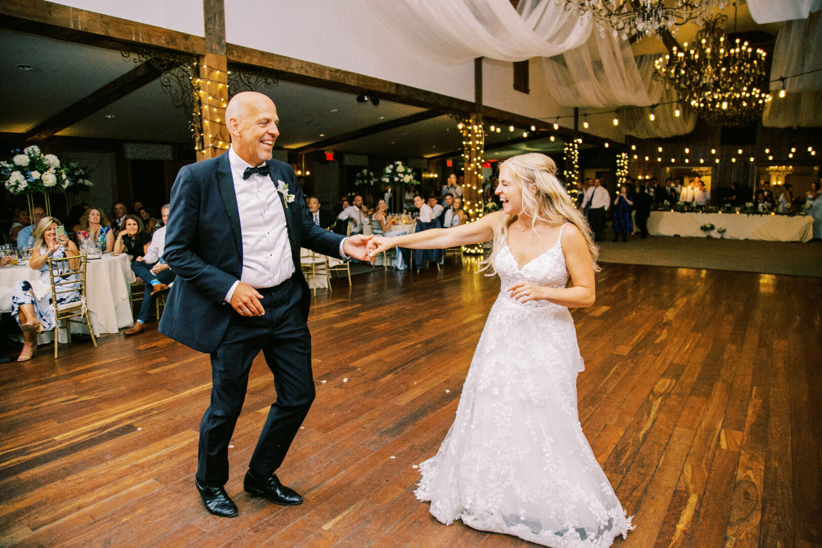 At a charming Normandy Farm wedding, a bride and an older man share a dance on the wooden floor, encircled by guests and twinkling string lights.