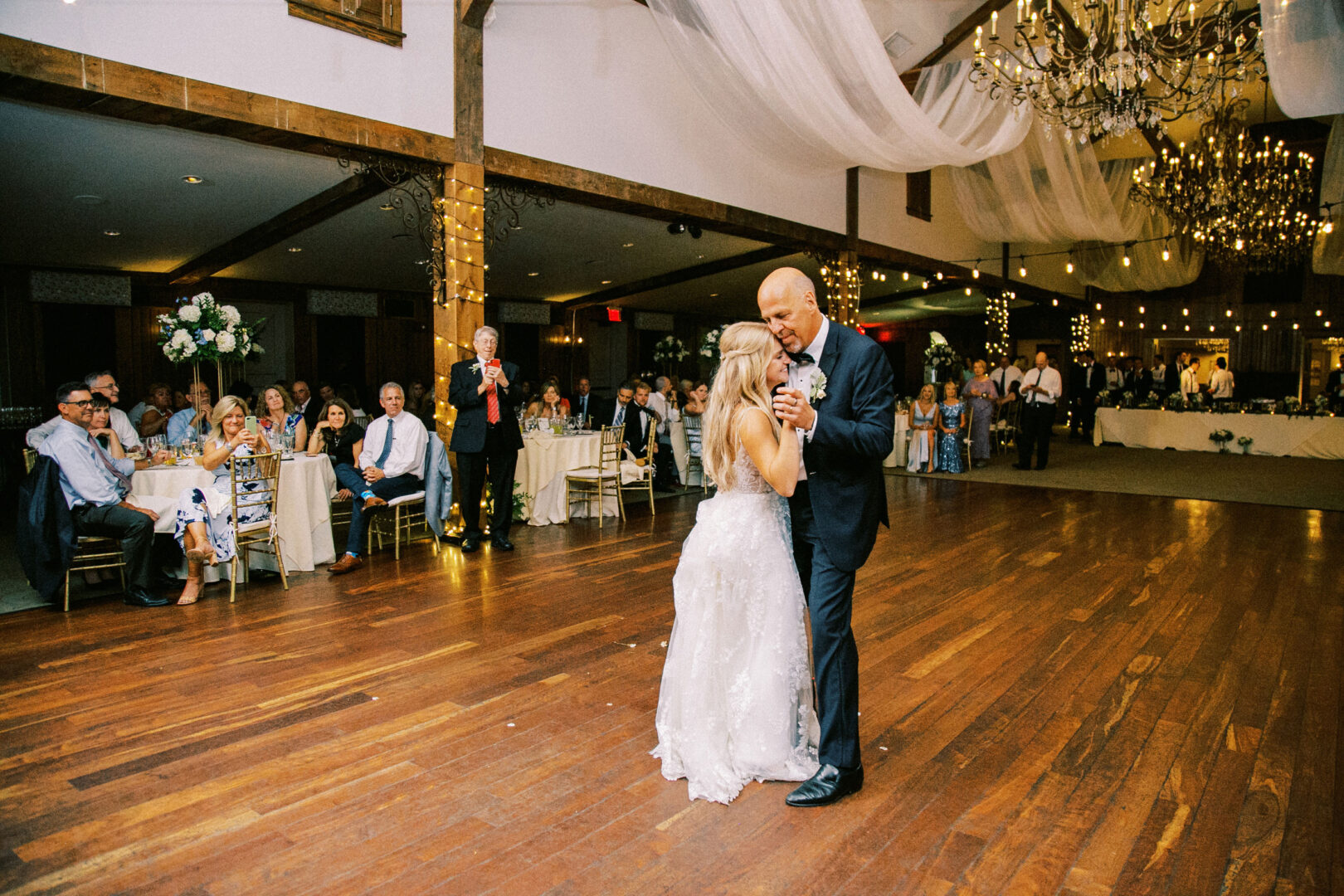 At a Normandy Farm wedding, a bride and an older man gracefully dance together in a large, elegantly decorated hall, as seated guests look on with admiration.