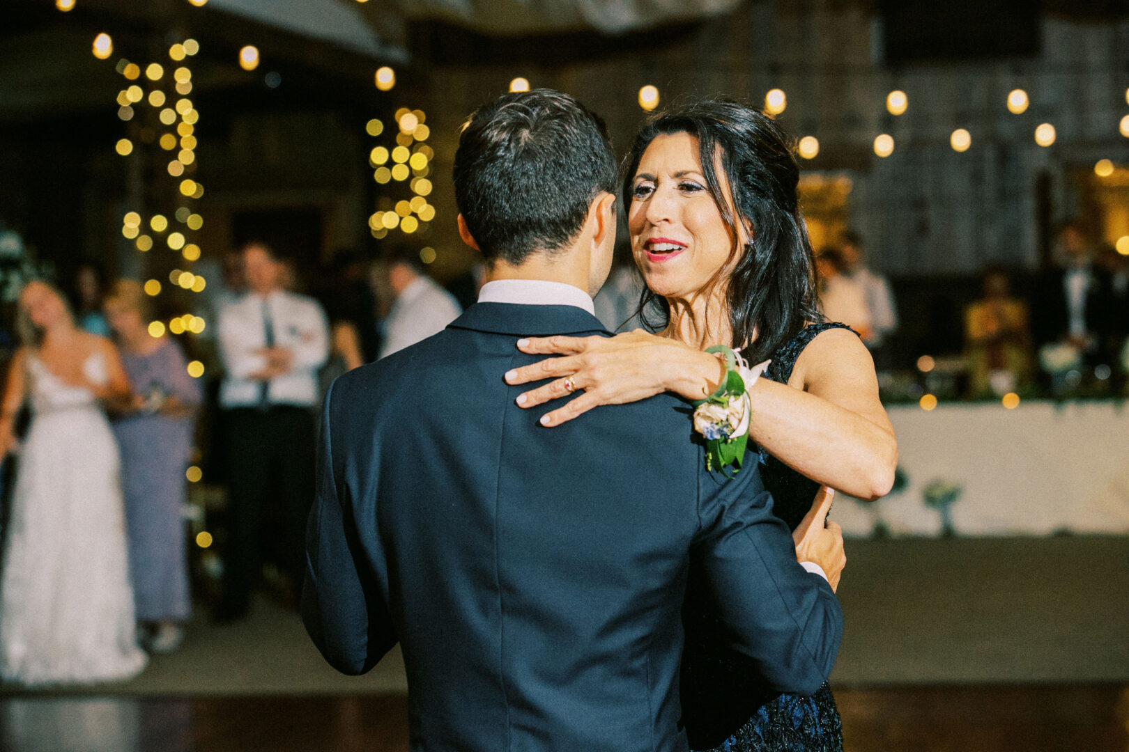 A woman and a man in formal attire are dancing closely at a Normandy Farm wedding, with string lights and elegantly decorated tables enhancing the atmosphere.