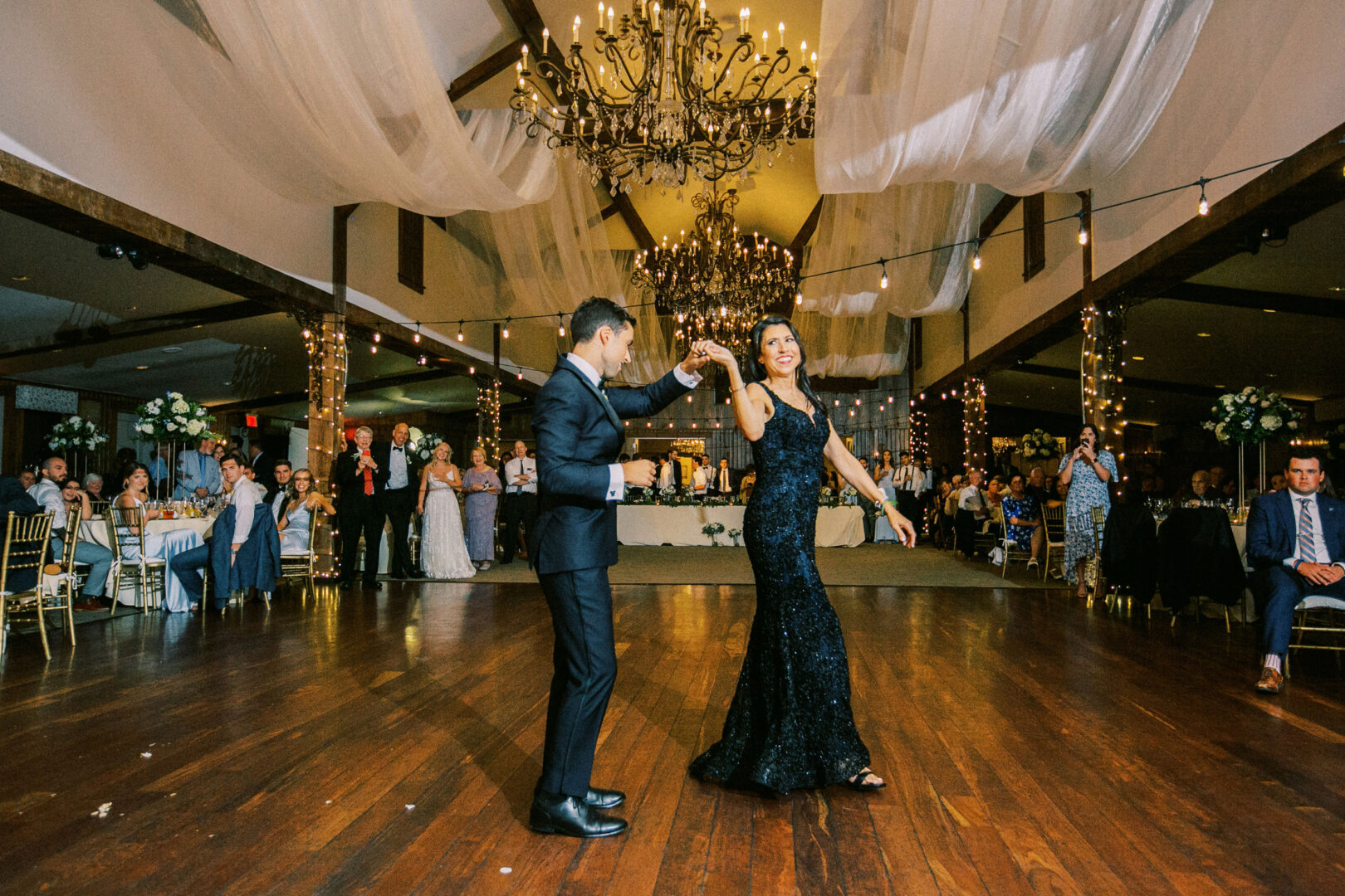 A couple dances under chandeliers in the elegant banquet hall of Normandy Farm, surrounded by guests seated at tables.