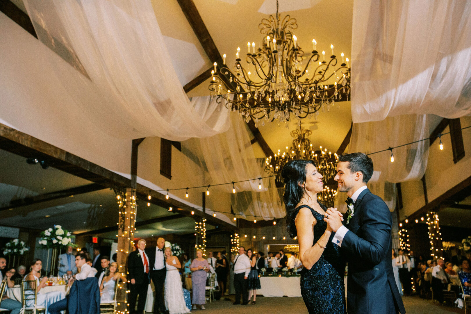 At a Normandy Farm Wedding, a couple dances in a decorated ballroom adorned with chandeliers and draped fabric. Guests watch from tables, while string lights illuminate the enchanting scene.