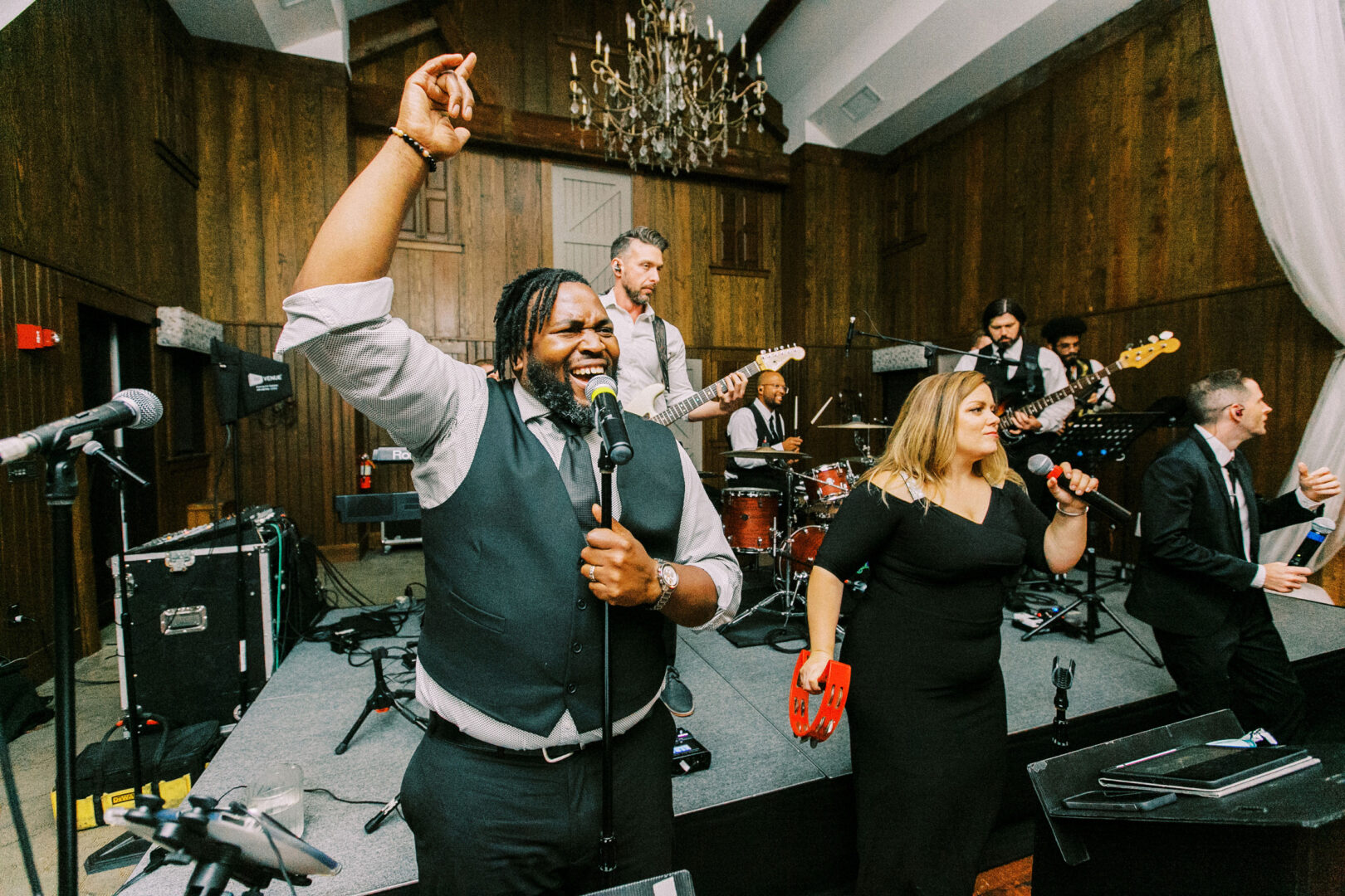 A band performs energetically on stage in a wooden-paneled room, reminiscent of a Normandy Farm Wedding. The vocalist raises an arm while a woman plays a tambourine, adding to the rustic charm, as musicians play instruments in the background.