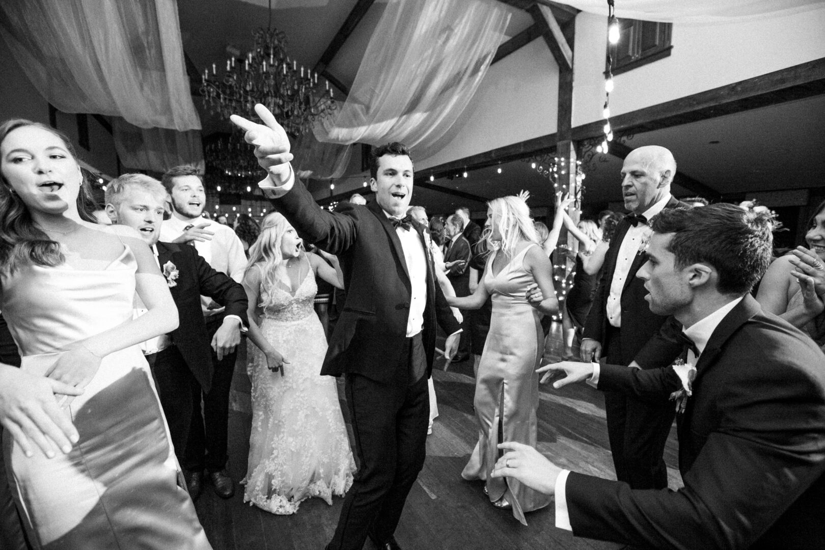 At a lively Normandy Farm wedding, a group in formal attire dances energetically beneath the chandelier and elegantly draped ceilings.