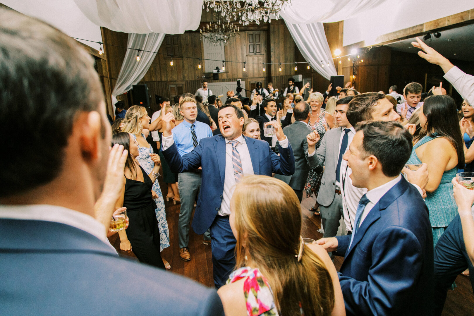 At a Normandy Farm wedding, people in formal attire dance energetically under a draped ceiling adorned with an elegant chandelier, creating a lively atmosphere at this crowded indoor event.