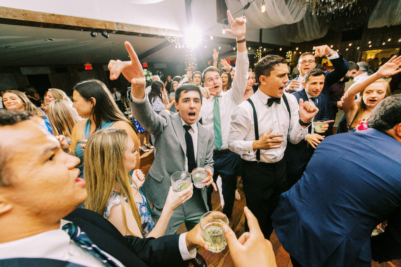 A lively crowd of well-dressed people dancing and holding drinks at an indoor celebration, reminiscent of a festive Normandy Farm wedding.