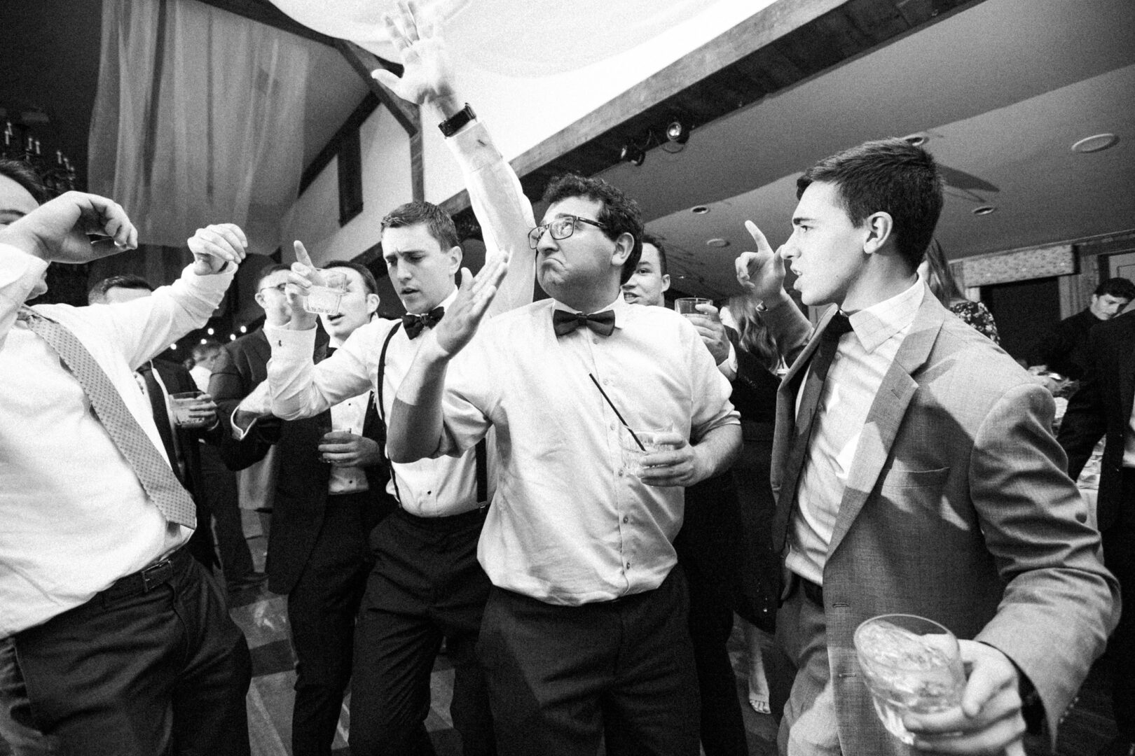 At a lively Normandy Farm Wedding, a group of men in formal attire energetically dance and gesture, adding excitement to the indoor social event.