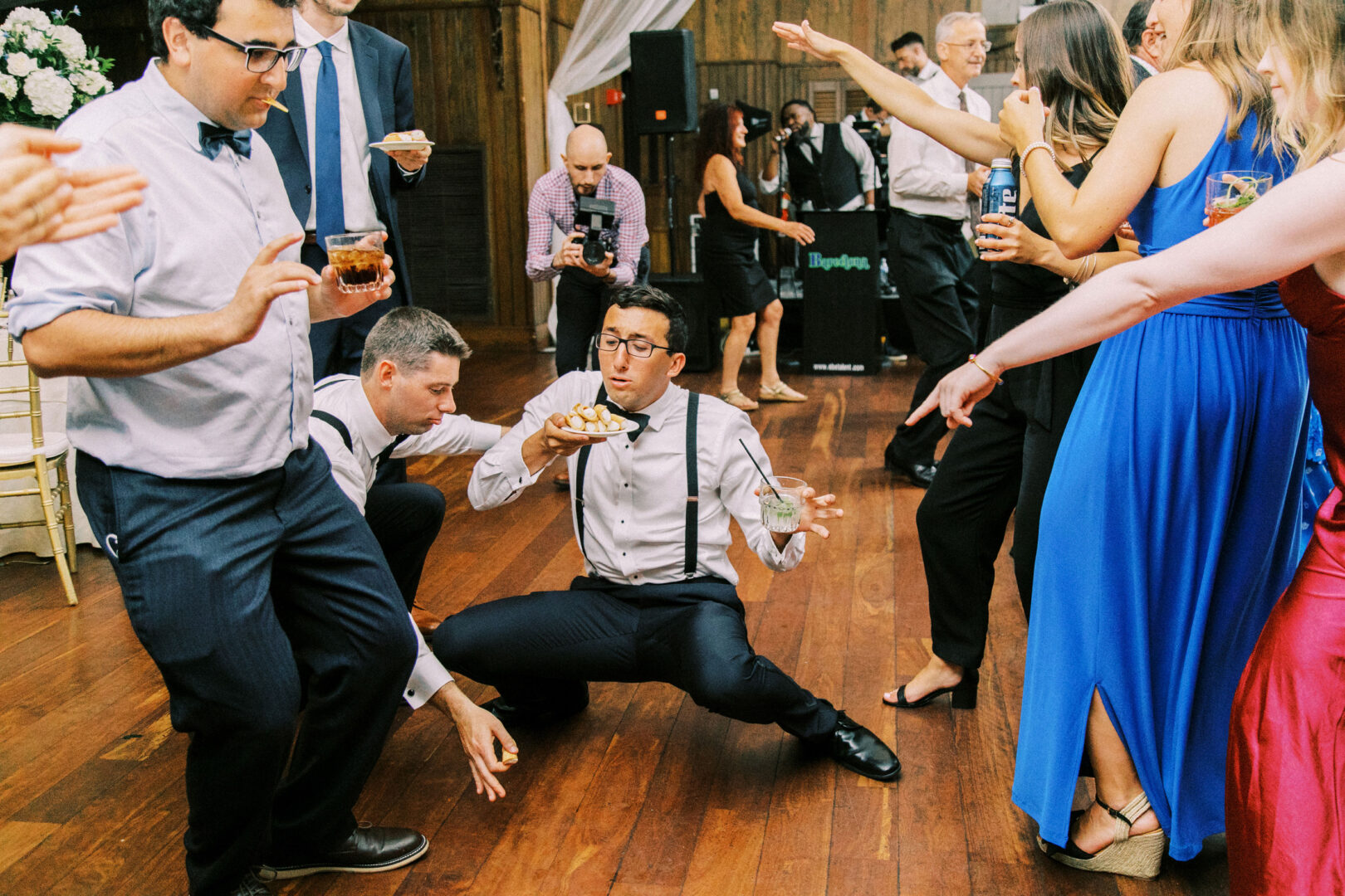 At a lively Normandy Farm wedding, a man in suspenders expertly balances a drink and a small plate on the dance floor, surrounded by guests dancing and pointing.