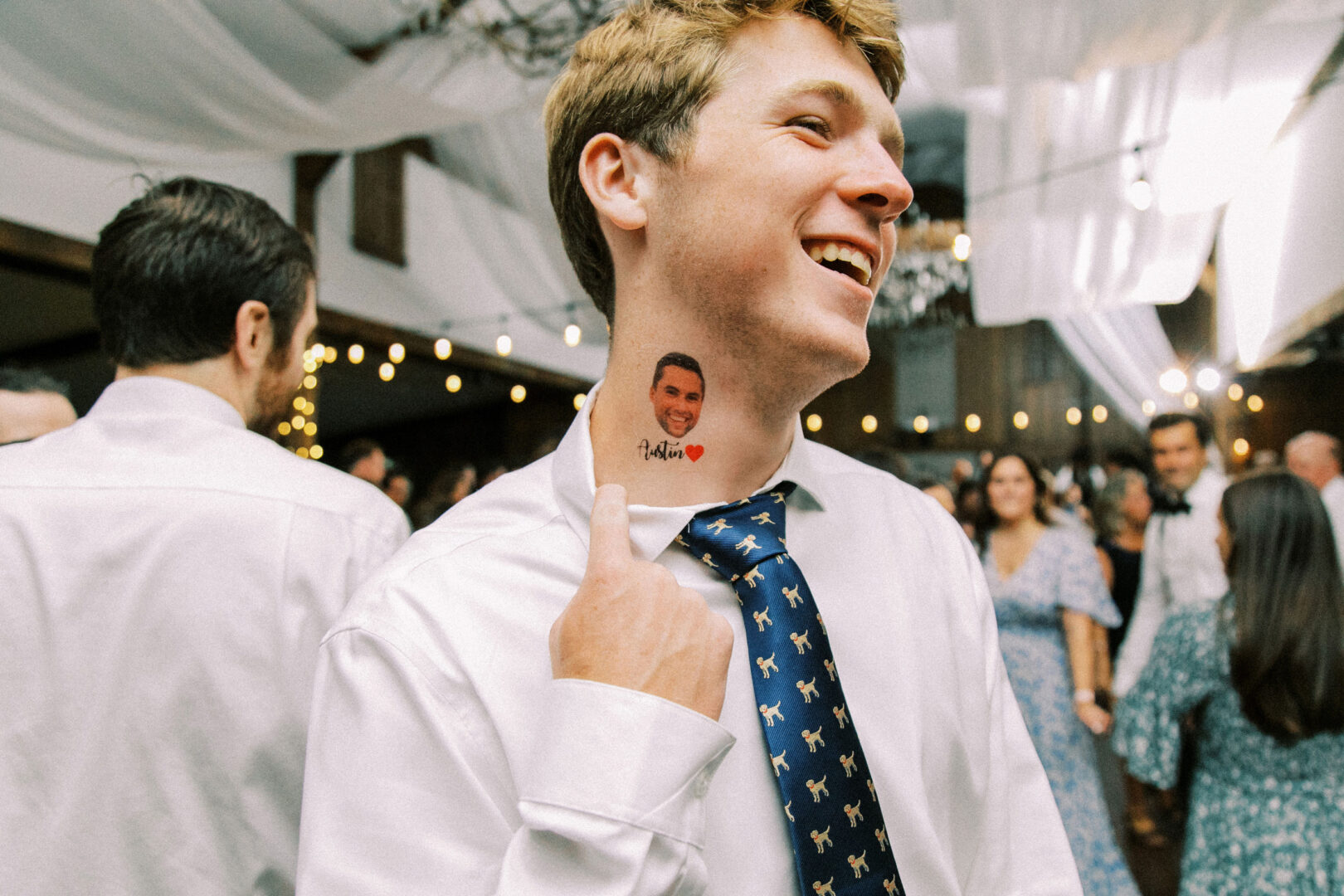 In a beautifully decorated indoor space reminiscent of a Normandy Farm wedding, a man in a white shirt and patterned tie smiles while pointing to a tattoo on his neck, surrounded by lively guests in the background.