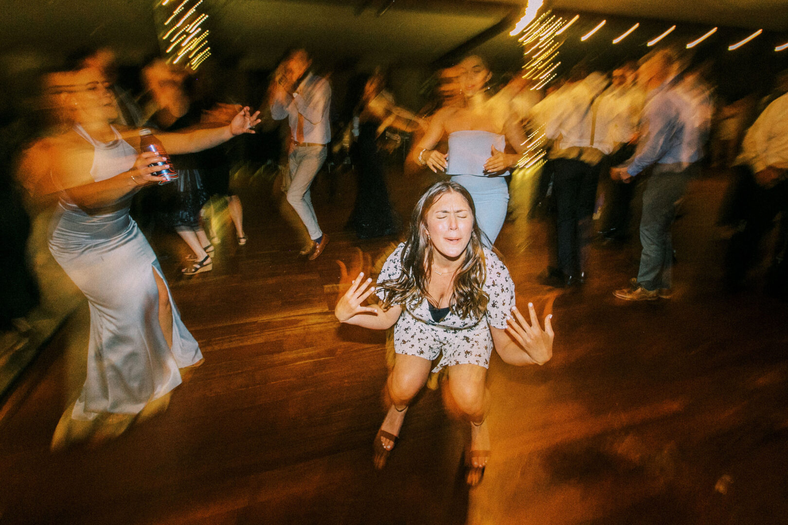 At a Normandy Farm Wedding, a woman in a dress dances energetically on the lively dance floor, with blurred people and twinkling lights creating an enchanting backdrop.