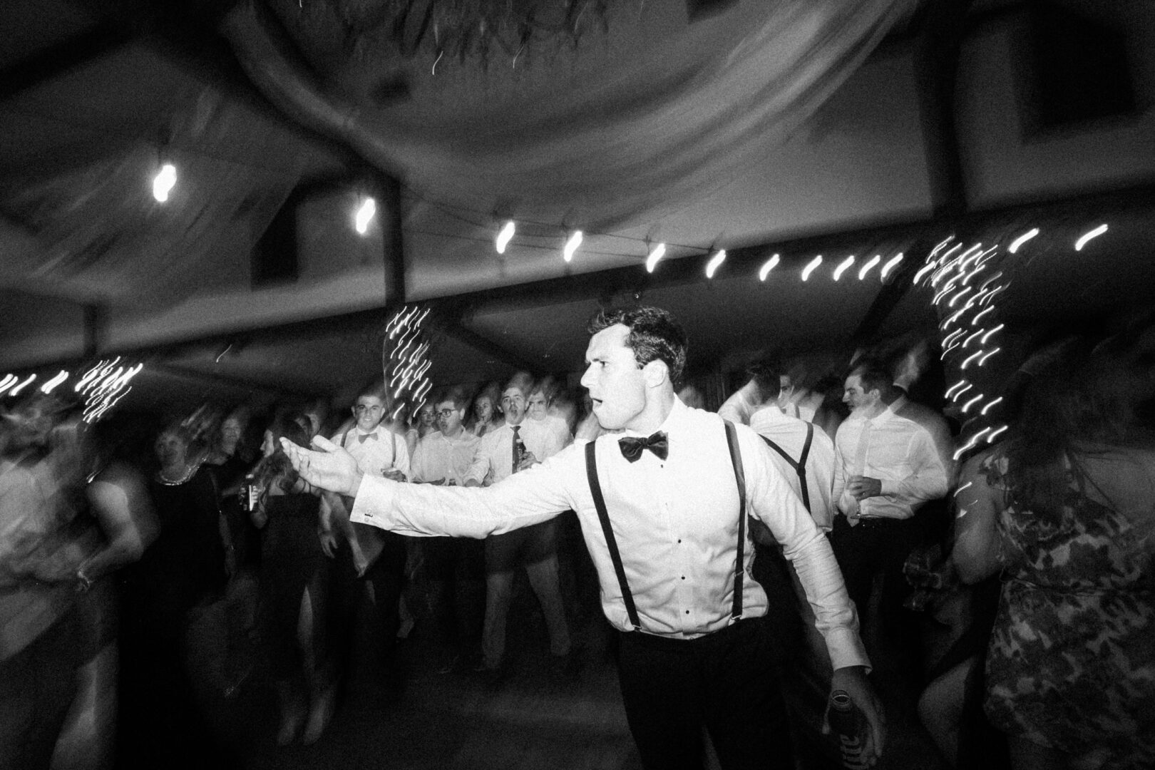 At an elegant Normandy Farm wedding, a man in formal attire energetically gestures on the crowded dance floor, setting the night's lively tone.