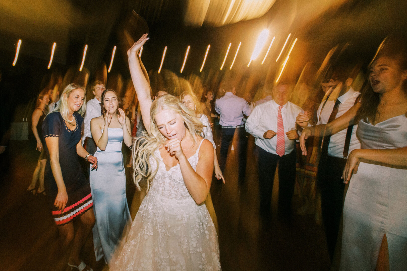 A bride in a white dress is dancing joyfully with a microphone at her Normandy Farm Wedding, surrounded by friends and guests at a lively reception.