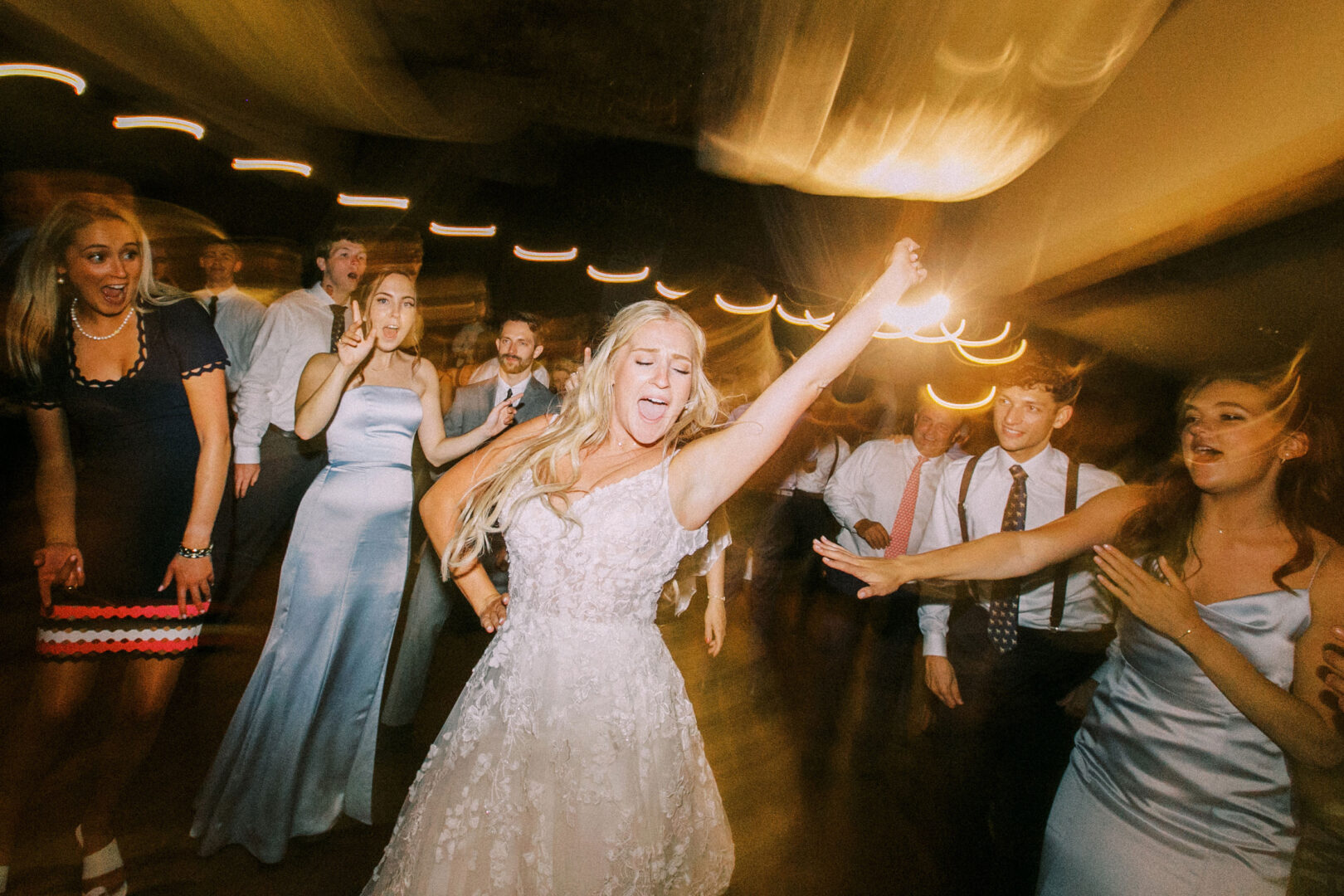 At a lively Normandy Farm wedding, the bride dances energetically on the dance floor, surrounded by cheering guests, with motion blur effects creating a vibrant backdrop.