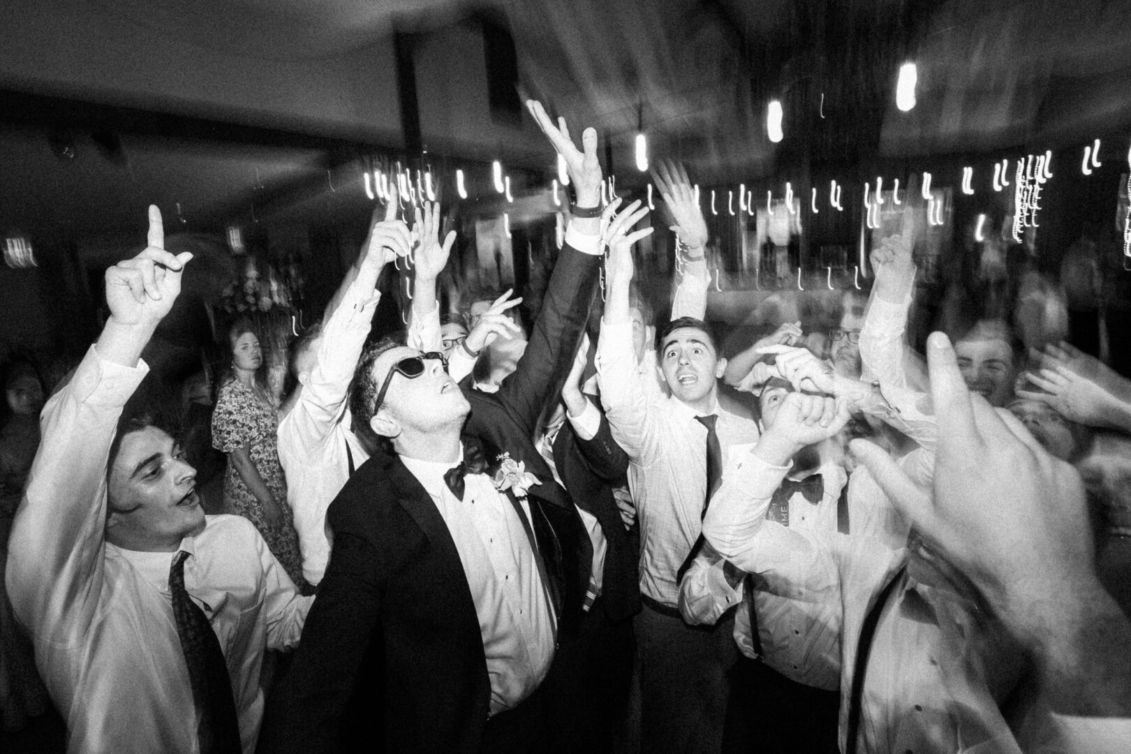 At a Normandy Farm wedding, a group of people in formal attire enthusiastically reach upward with their hands at an indoor event.