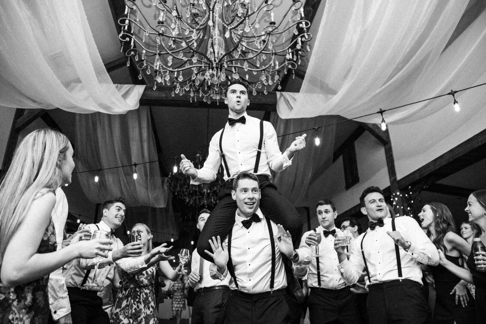 At a vibrant celebration reminiscent of a Normandy Farm wedding, a man in a tuxedo rides on another's shoulders amidst a lively crowd gathered under the glowing chandelier.