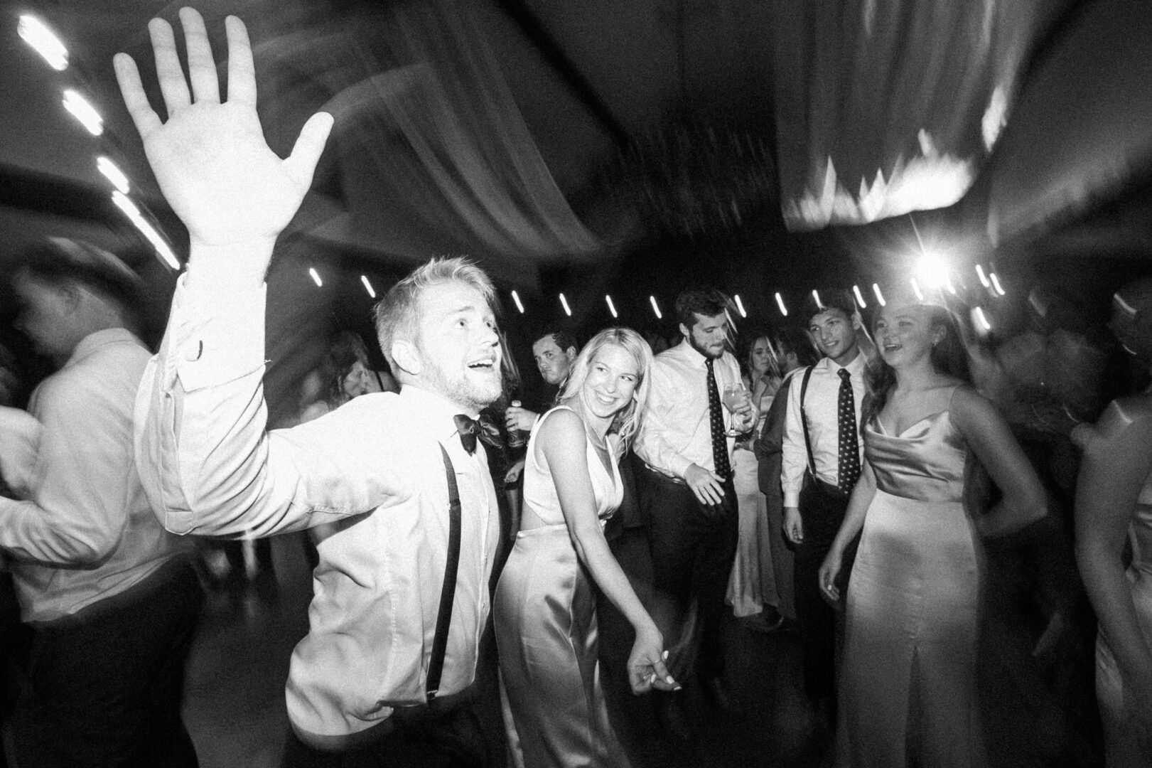 At a stunning Normandy Farm wedding, a group of people in formal attire dance energetically at the party. One person in the foreground raises a hand, while others smile and enjoy the lively atmosphere.