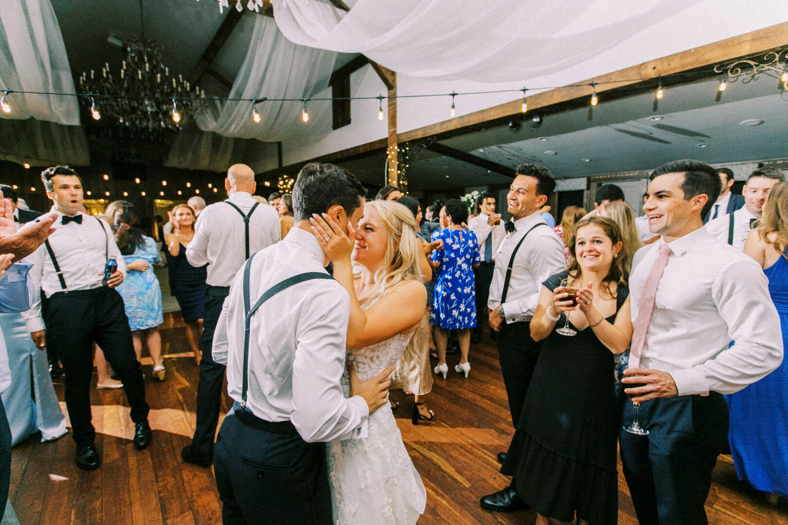 At a Normandy Farm wedding, a couple embraces on the dance floor, surrounded by smiling guests. The venue's rustic charm is evident with its wooden floors and twinkling string lights.