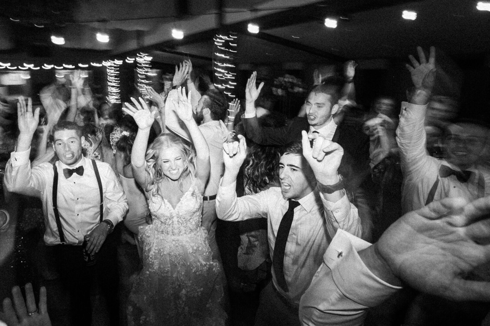 Black and white photo of a lively Normandy Farm wedding celebration. A group of people, including the bride and groom, are dancing with hands raised in the air.