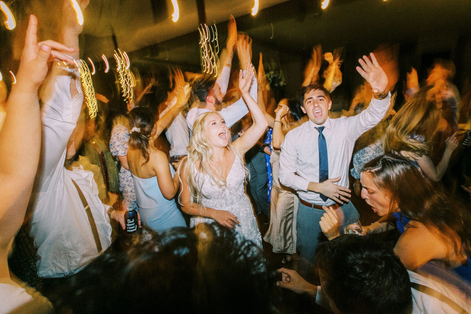 A joyful Normandy Farm wedding reception unfolds as the bride and groom dance amid guests. The lively crowd, with hands raised in celebration, mirrors the enchanting atmosphere of the venue.