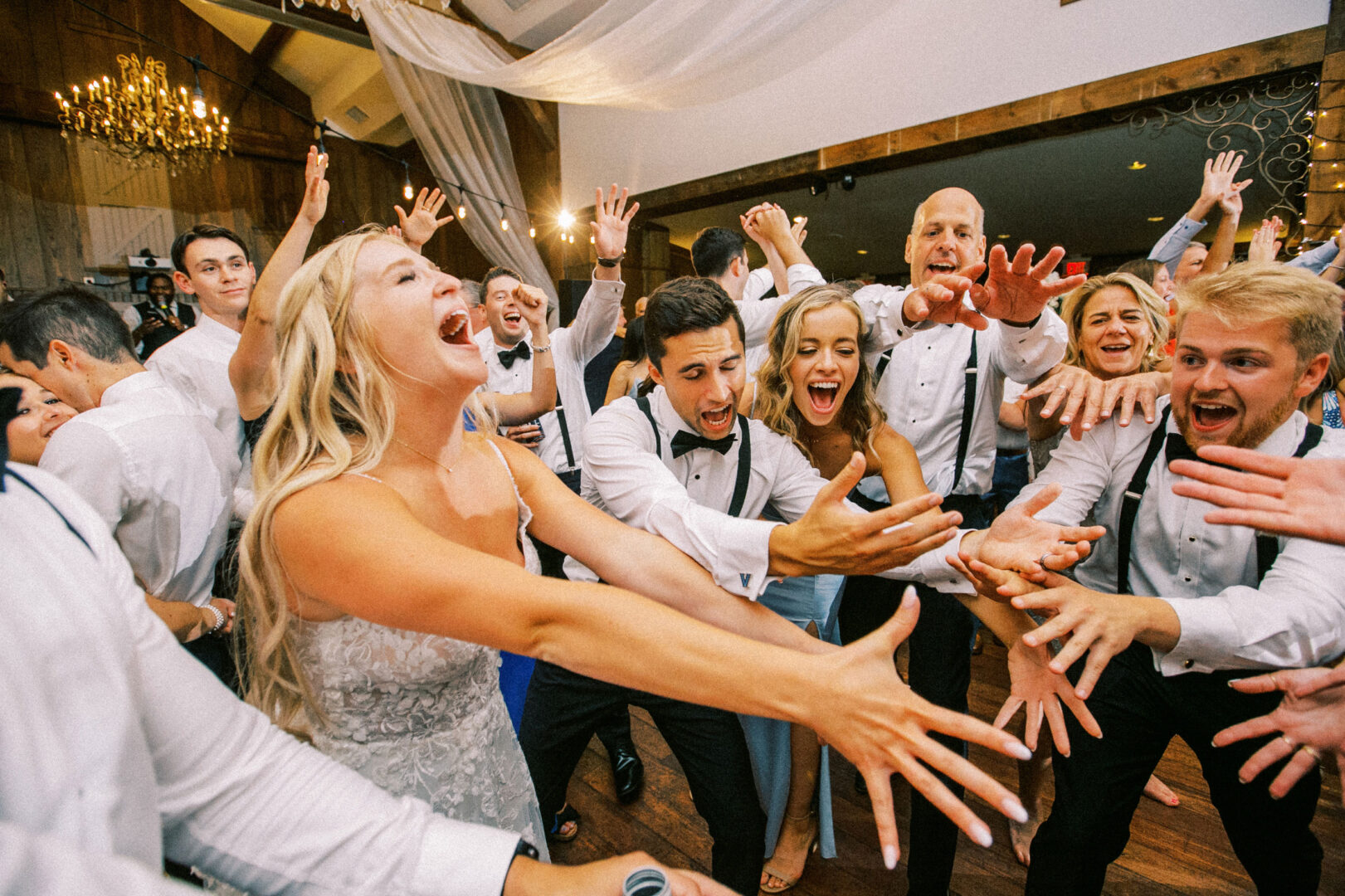 At a Normandy Farm wedding, a group of people, including the bride, joyfully reach their hands toward the center at a celebration in a beautifully decorated hall.