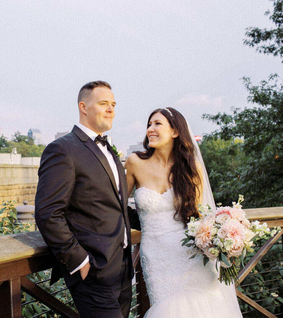 A couple in wedding attire stands on a wooden walkway at Waterworks. The groom wears a suit, and the bride holds a bouquet, framed by a cityscape and trees in the background.