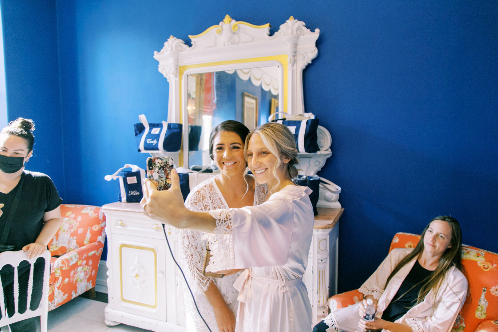Two women in robes take a selfie in a room with blue walls and a decorative mirror. Others sit nearby, one wearing a face mask, next to gifts on a dresser.