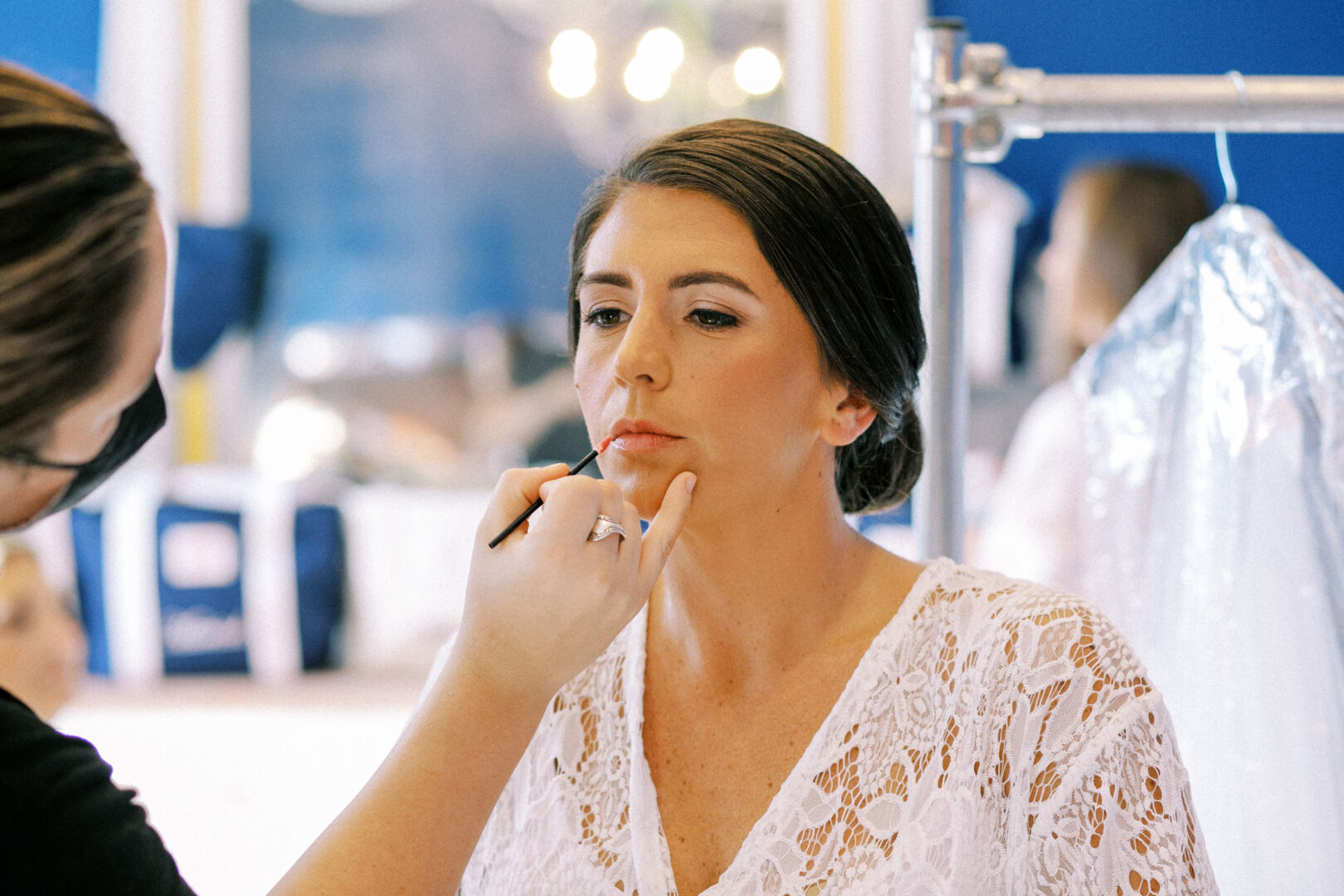A woman in a white lace top is having makeup applied by a makeup artist in a well-lit room.