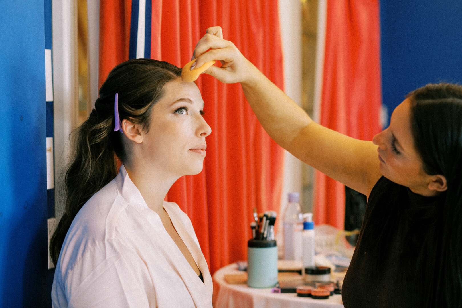 A woman applies makeup to another woman's forehead with an orange sponge in a room with red curtains and a table with makeup tools.