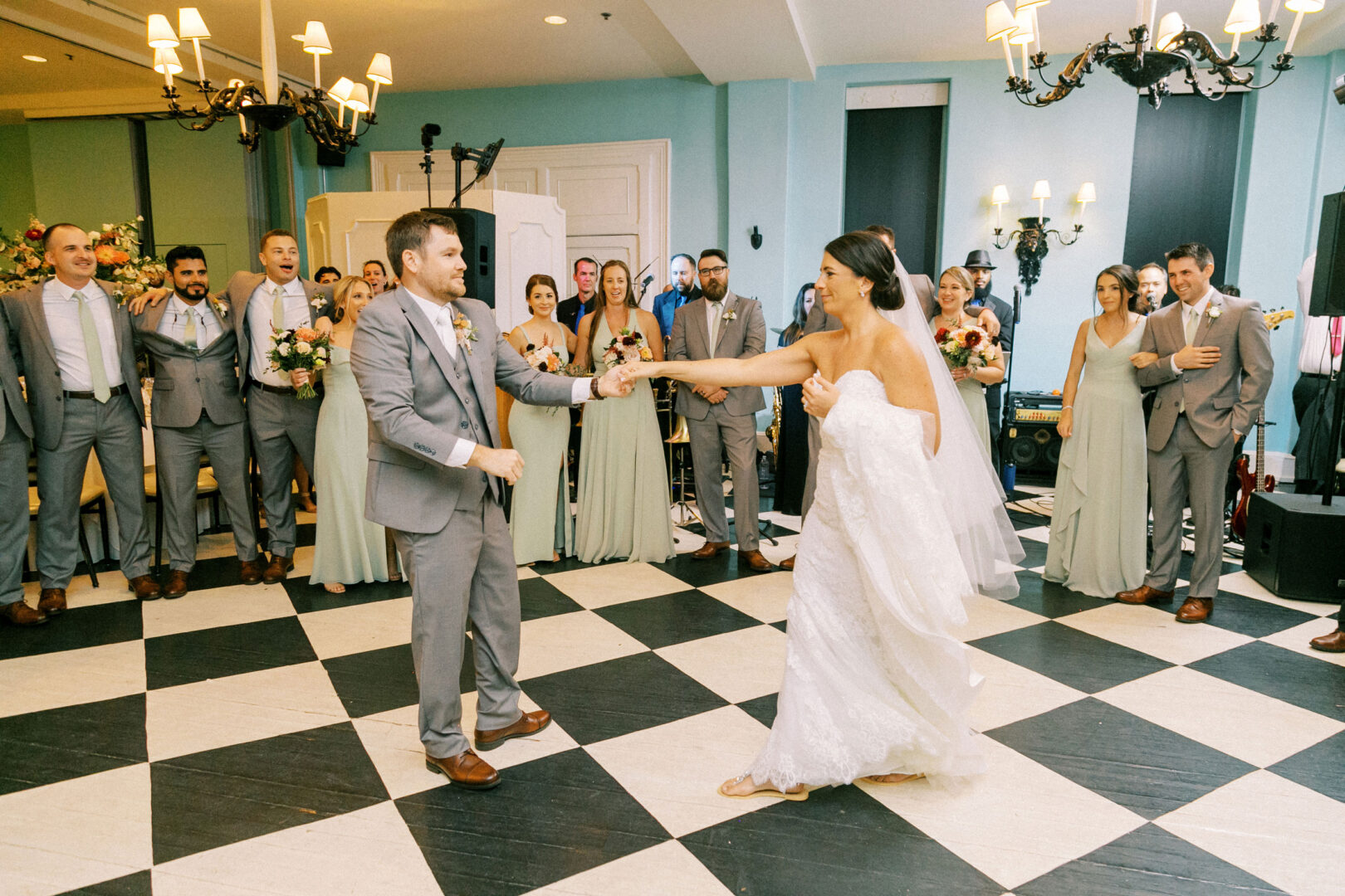 A bride and groom dance on a black-and-white checkered floor. Bridesmaids and groomsmen watch in the background. The room has teal walls and chandeliers.