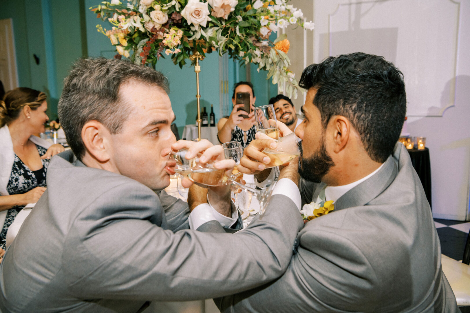 Two men in suits are linked arm-in-arm, drinking from champagne glasses at a celebration. A person in the background takes a photo with a phone. There is a floral arrangement behind them.