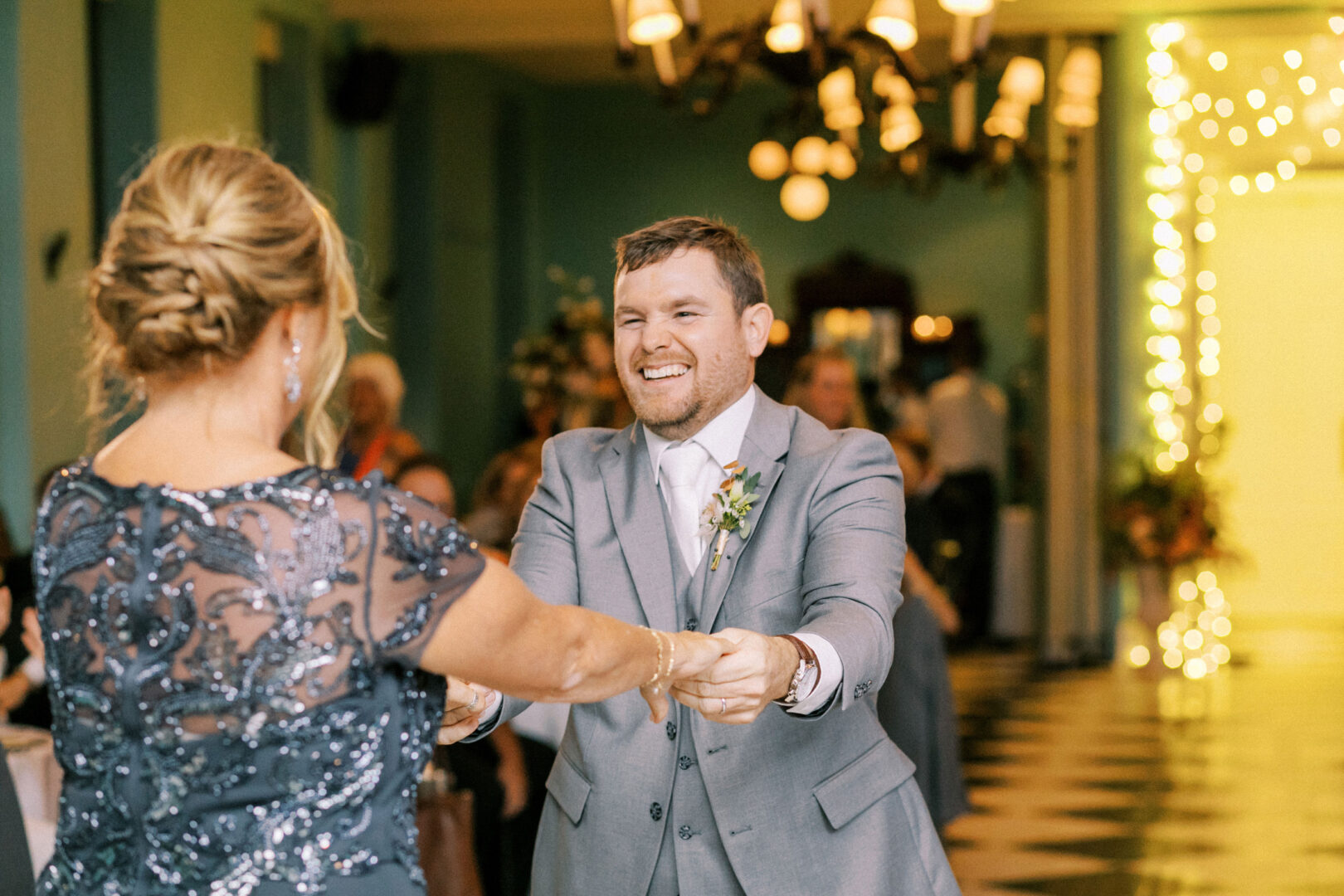 A man and woman dance together in a warmly lit room with checkered flooring and chandeliers. The man is smiling and wearing a gray suit, and the woman is in a sequined dress.