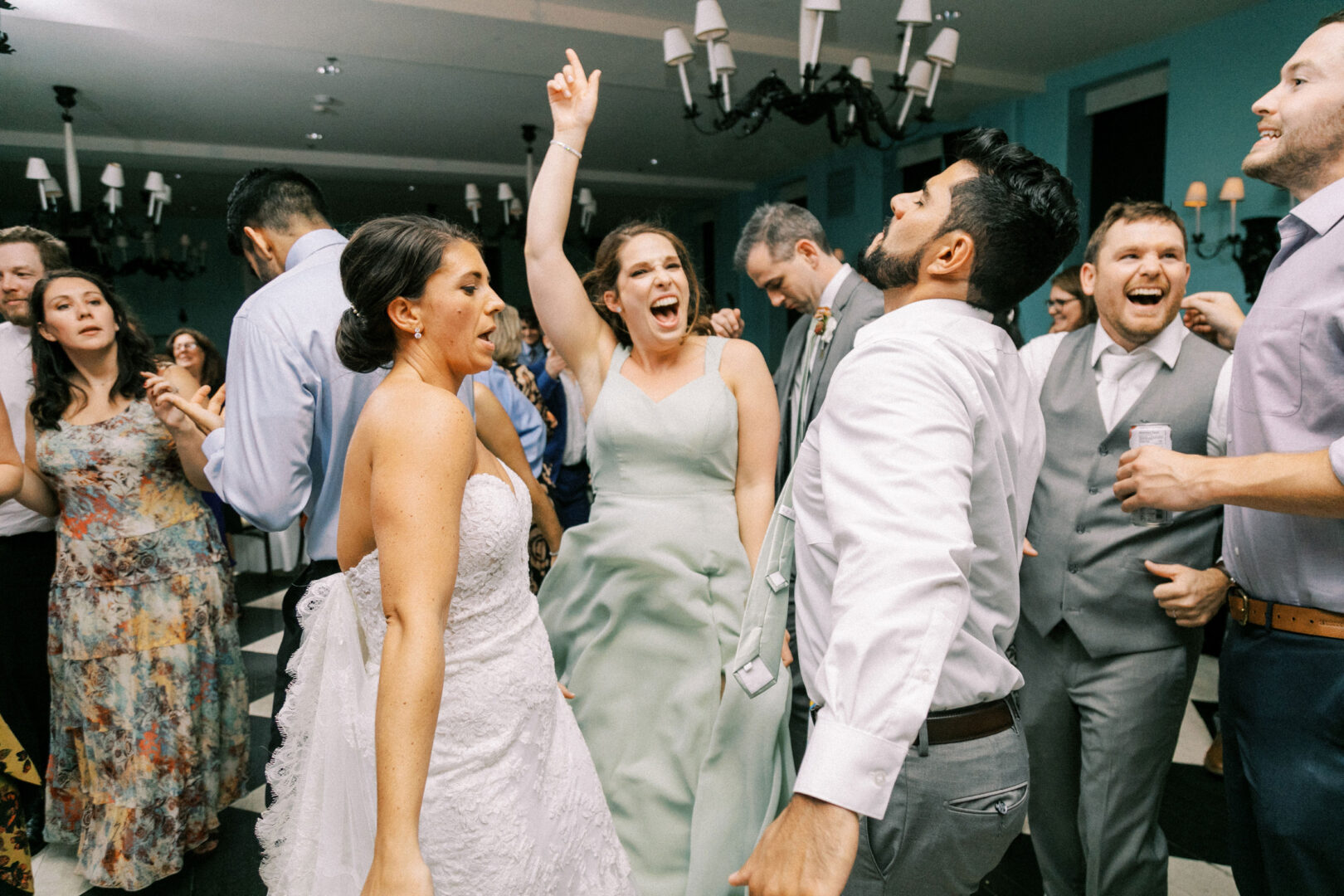 Group of people in formal attire dancing energetically at an indoor event. A woman in a white dress and a woman in a green dress are in the foreground. Others are smiling and dancing around them.