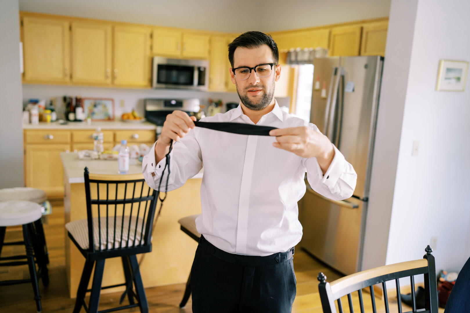 A man stands in a kitchen, dressed in a crisp white shirt and holding a tie, preparing for a Cape May wedding.