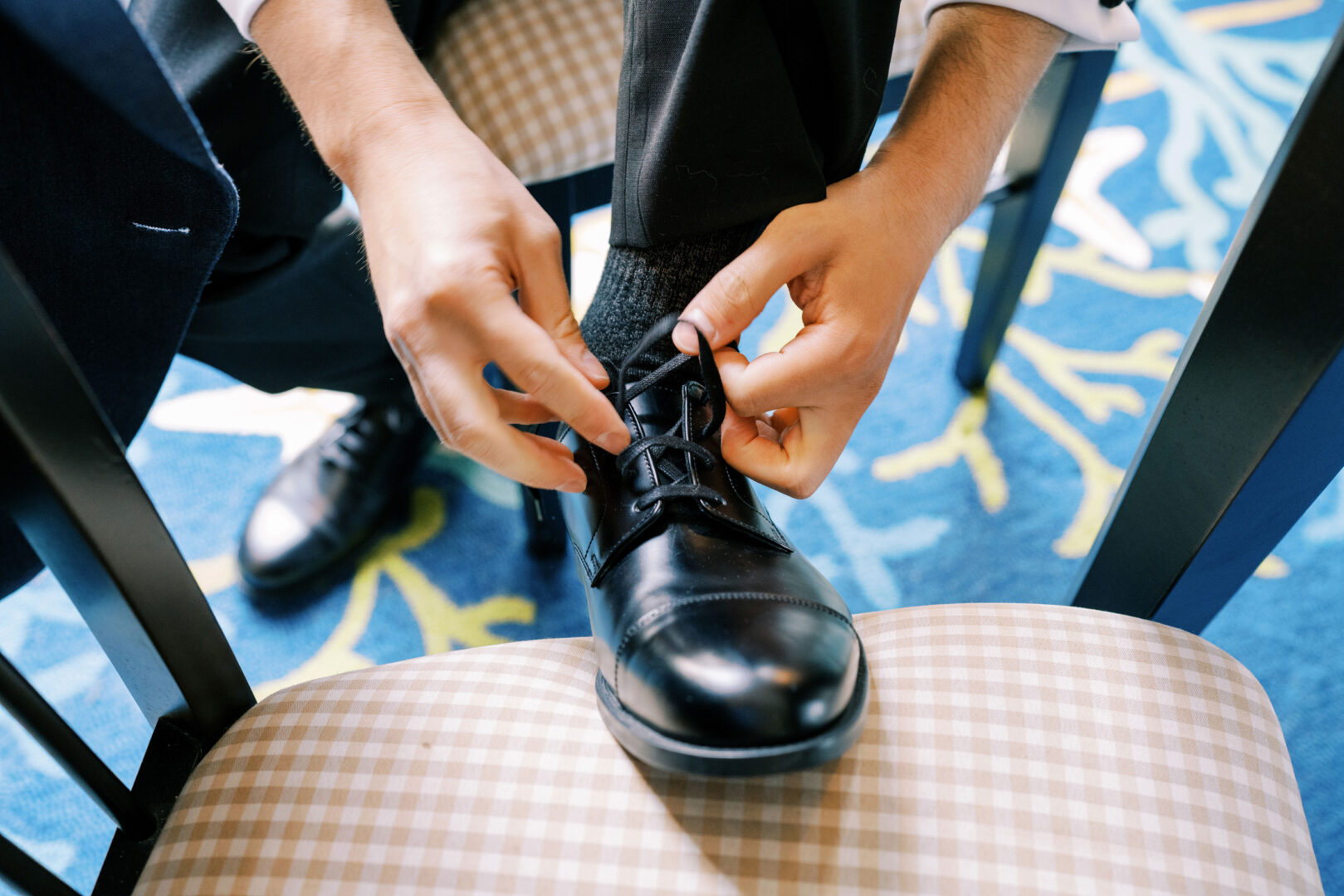 Seated on a chair with a blue carpet pattern beneath, a person meticulously ties the shoelaces on their black formal shoes, preparing for the elegant Cape May wedding.