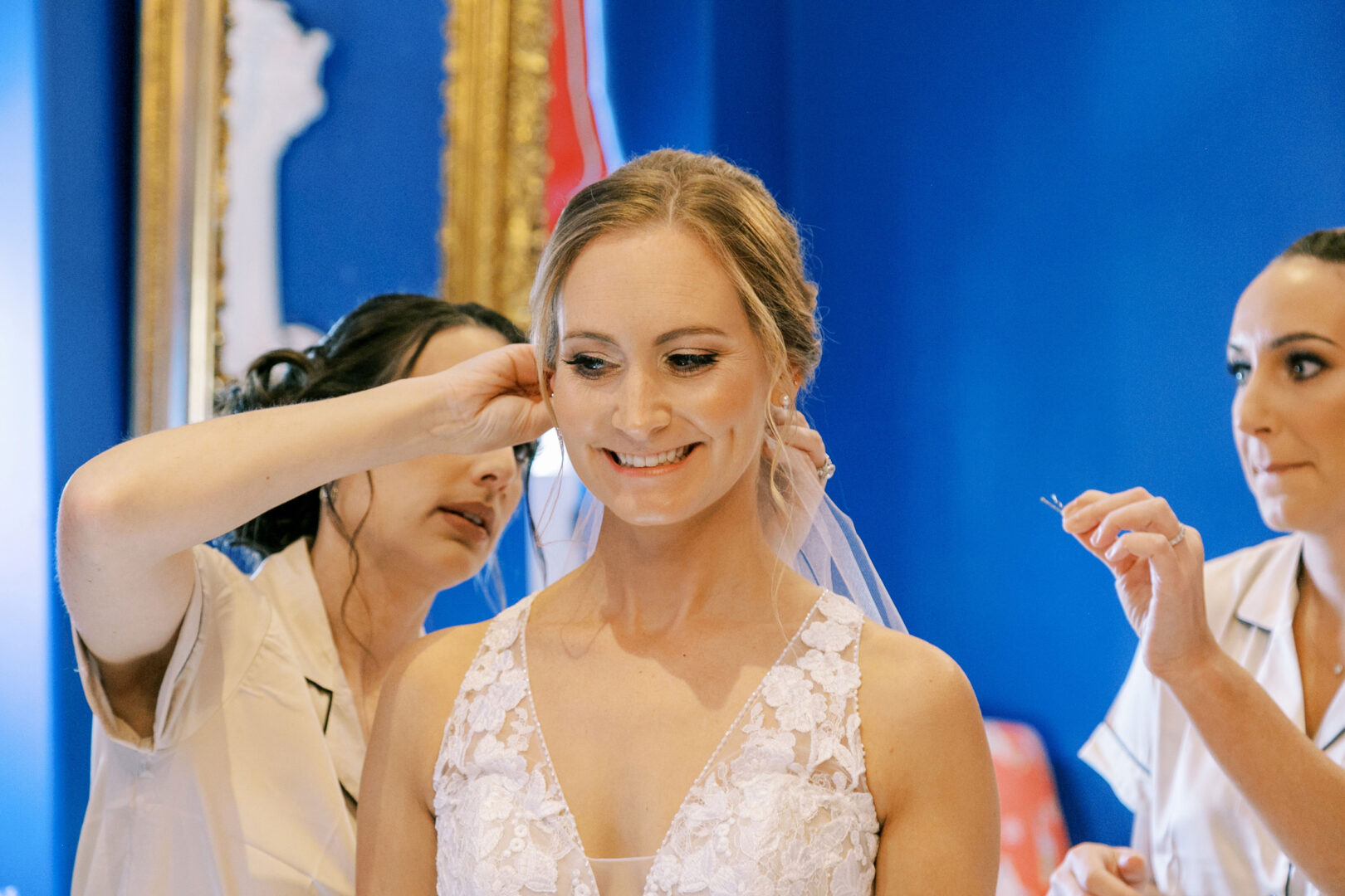 Bride smiling as two women adjust her veil in a room with blue walls and a gold-framed mirror, capturing the elegance of a Cape May wedding.