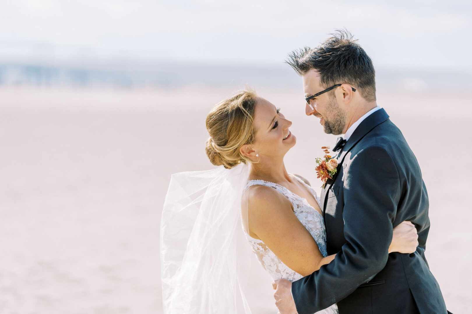 A bride and groom embrace on a Cape May beach. The bride wears a white dress and veil, the groom in a black suit with a boutonniere. The sandy background and clear sky add to the enchanting atmosphere of their Cape May wedding.