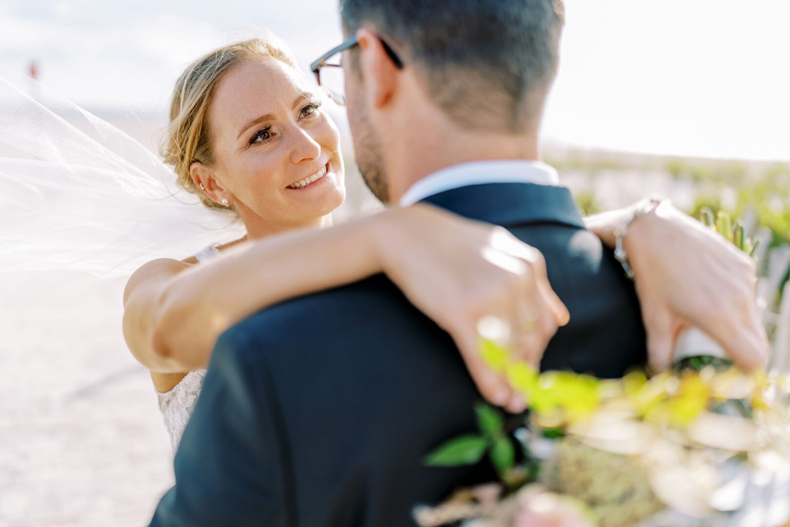 At a charming Cape May wedding, a bride beams as she embraces her groom under the open sky, his arms gently cradling a vibrant bouquet.