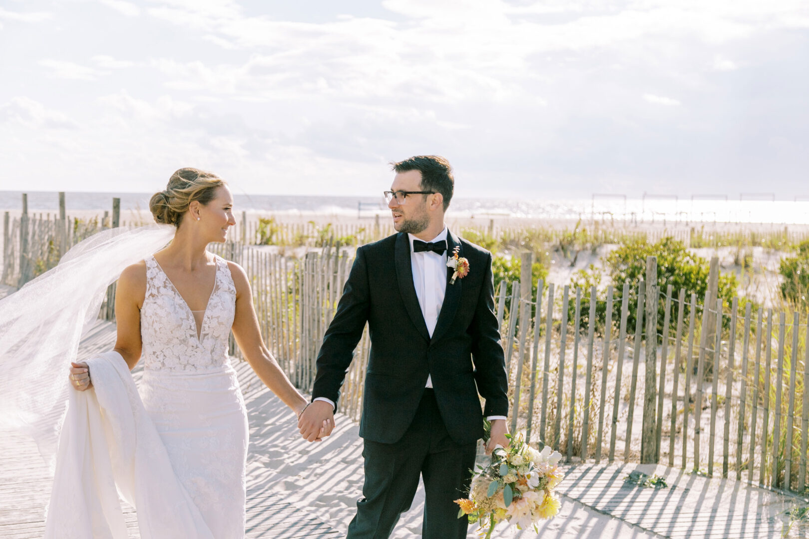 A bride and groom walk hand in hand on a Cape May beach, with sand dunes and a fence in the background. The groom is dashing in his black suit, while the bride dazzles in her white dress and veil, holding a bouquet.