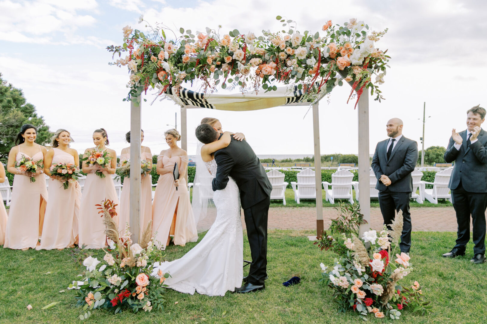 At a Cape May wedding, a couple kisses under a floral arch during the outdoor ceremony. Bridesmaids in peach dresses and groomsmen in suits stand nearby, completing the romantic scene.