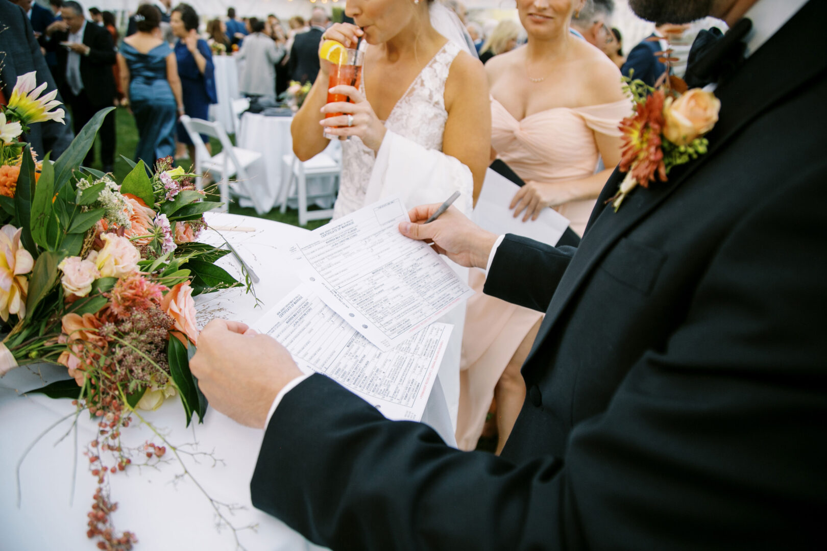 At a charming Cape May wedding, a guest reviews documents at the reception, looking sharp in a suit with a boutonnière. Surrounded by elegant floral arrangements, other attendees in formal attire bustle about in the background, adding to the festive ambiance.