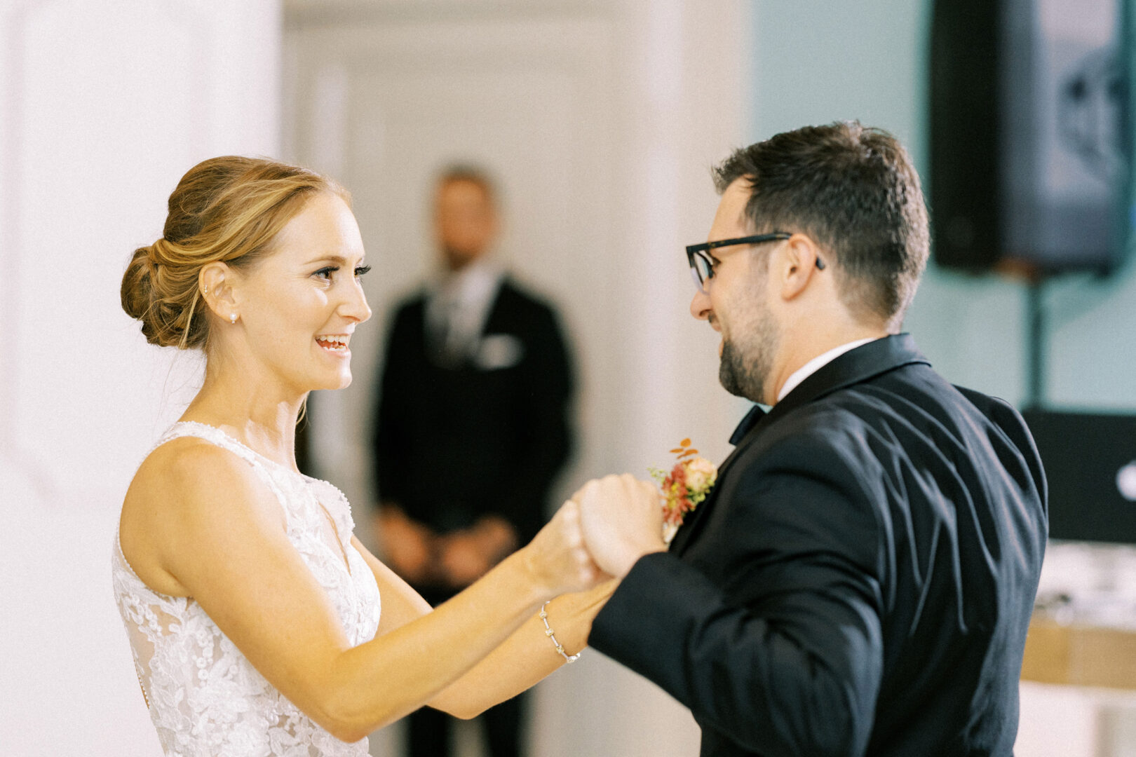 At a charming Cape May wedding, a bride and groom dance together, smiling in the warmly lit room, as a man in a suit stands gracefully in the background.