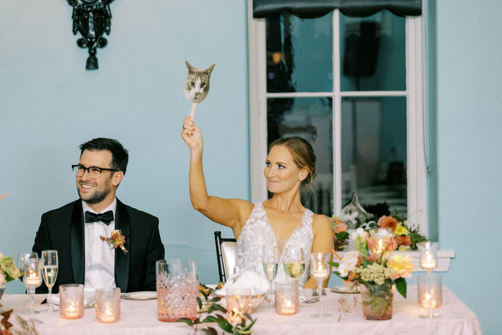 At their enchanting Cape May wedding, the bride playfully holds a cat face sign beside her beaming groom at a beautifully decorated table adorned with candles and flowers.