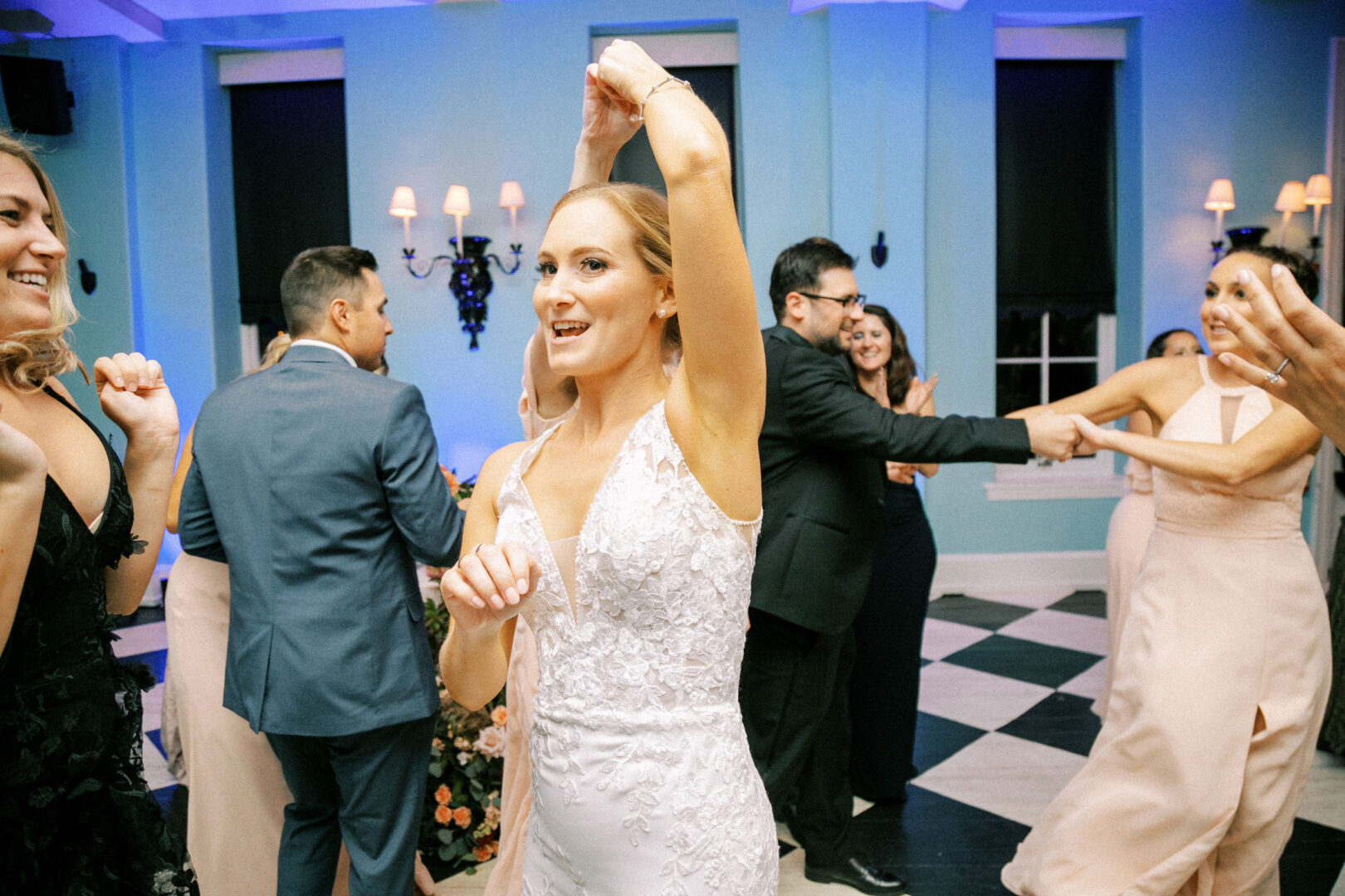 At a lively Cape May wedding, people dance energetically across a checkered floor beneath blue walls. A woman in a white dress stands at the center, joyfully surrounded by celebrants who share in her happiness and revelry.