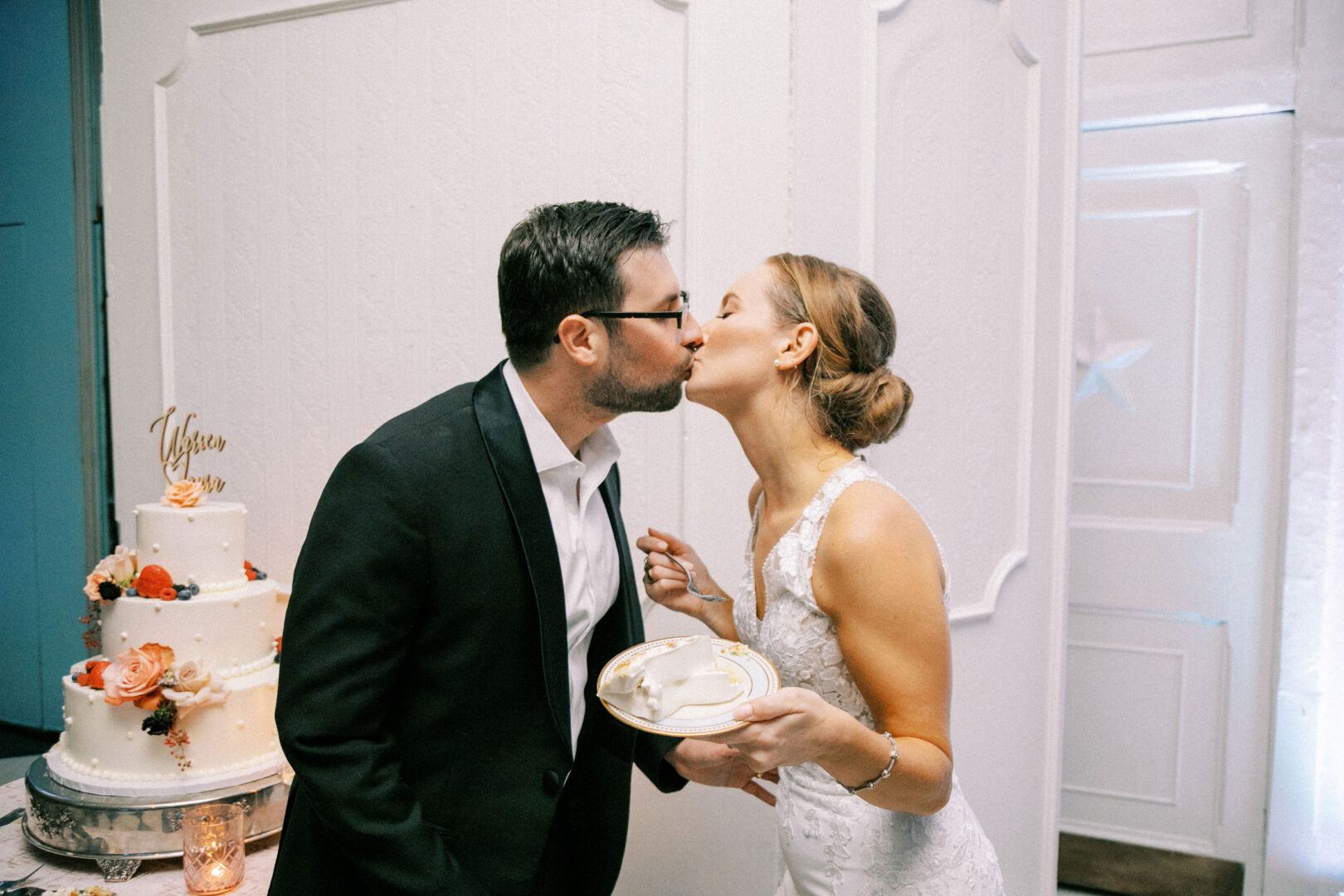 A couple kisses in front of a white wedding cake at their Cape May wedding. The groom wears a black suit, and the bride, in her beautiful white dress, holds a plate with cake.