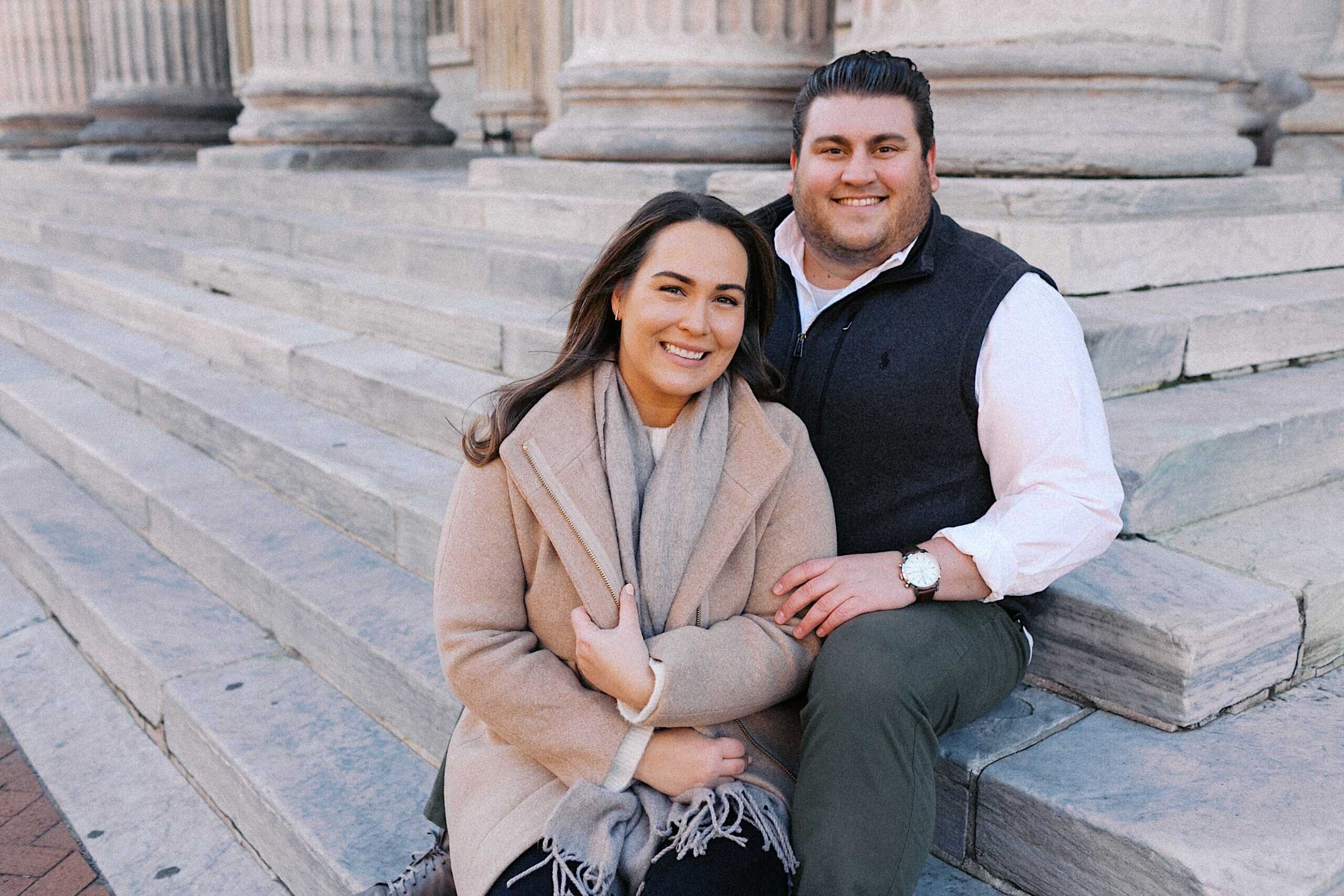 A woman and a man sit on stone steps in front of large columns, both smiling at the camera.
