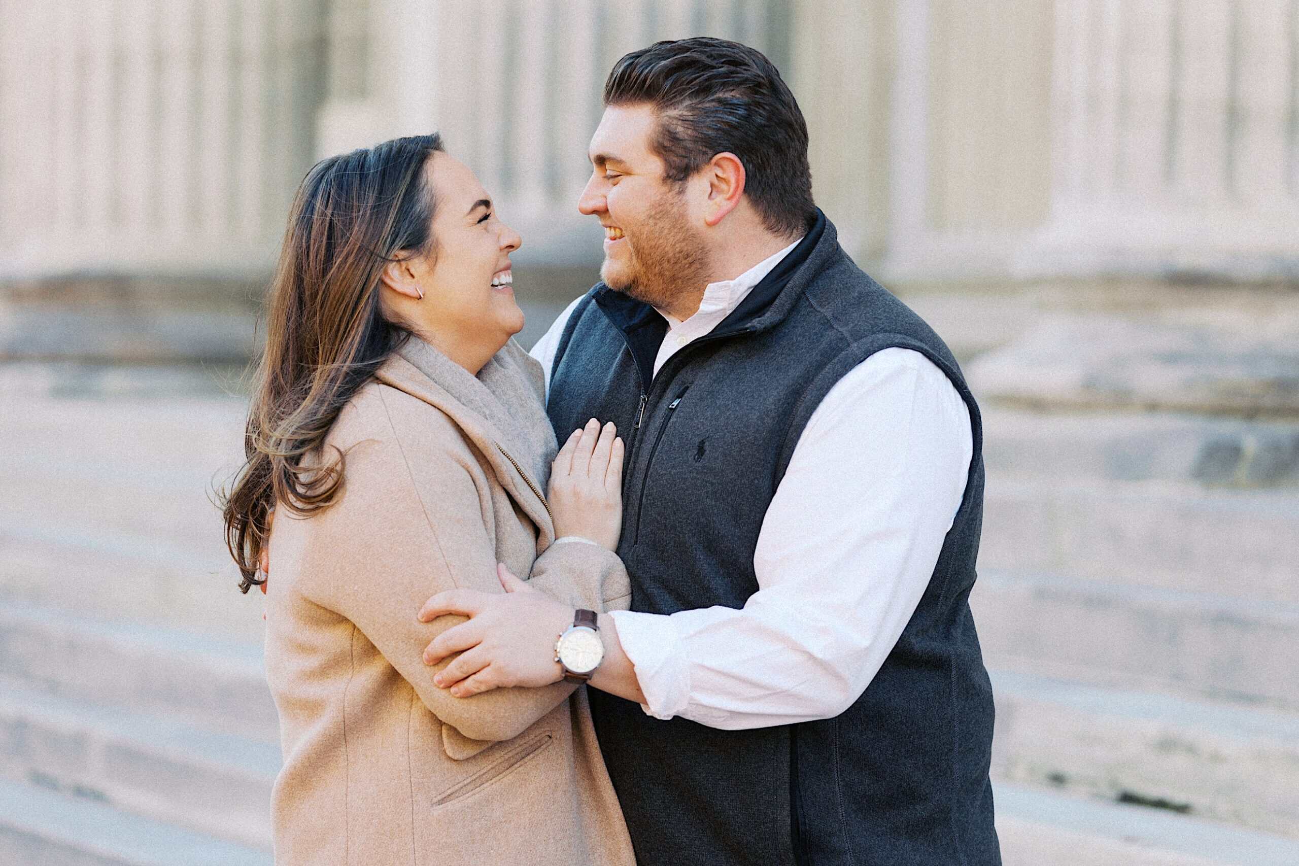 A man and woman stand close together outdoors, smiling at each other on stone steps in front of a building with columns.